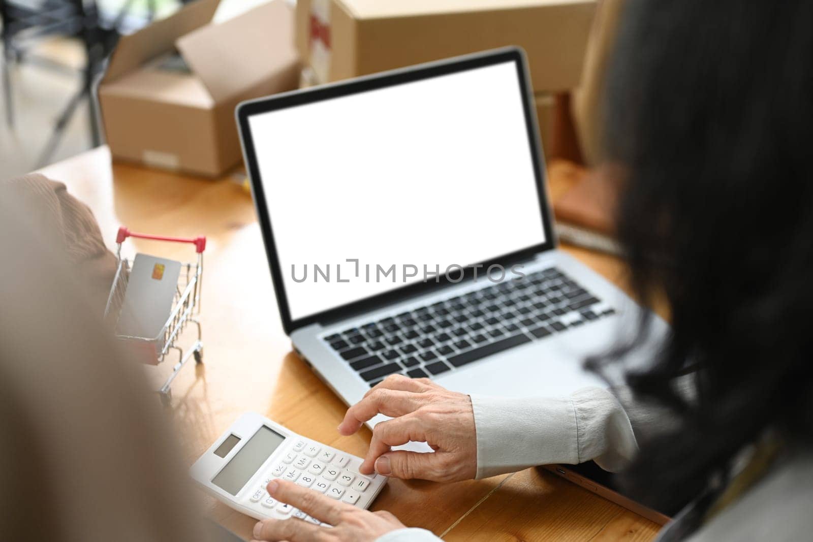 Small business owner sitting in front of laptop with blank screen and calculating expenses.