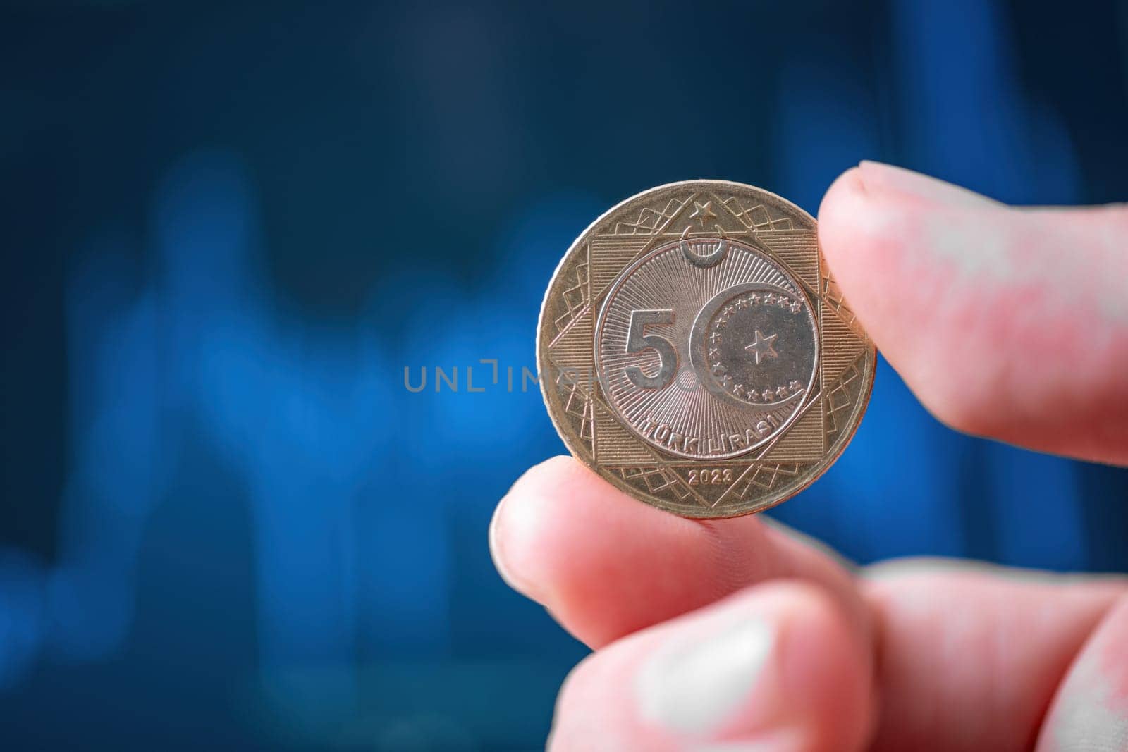 Hand holding a 5 Turkish Lira coin in front of a screen with financial chart curves by Sonat