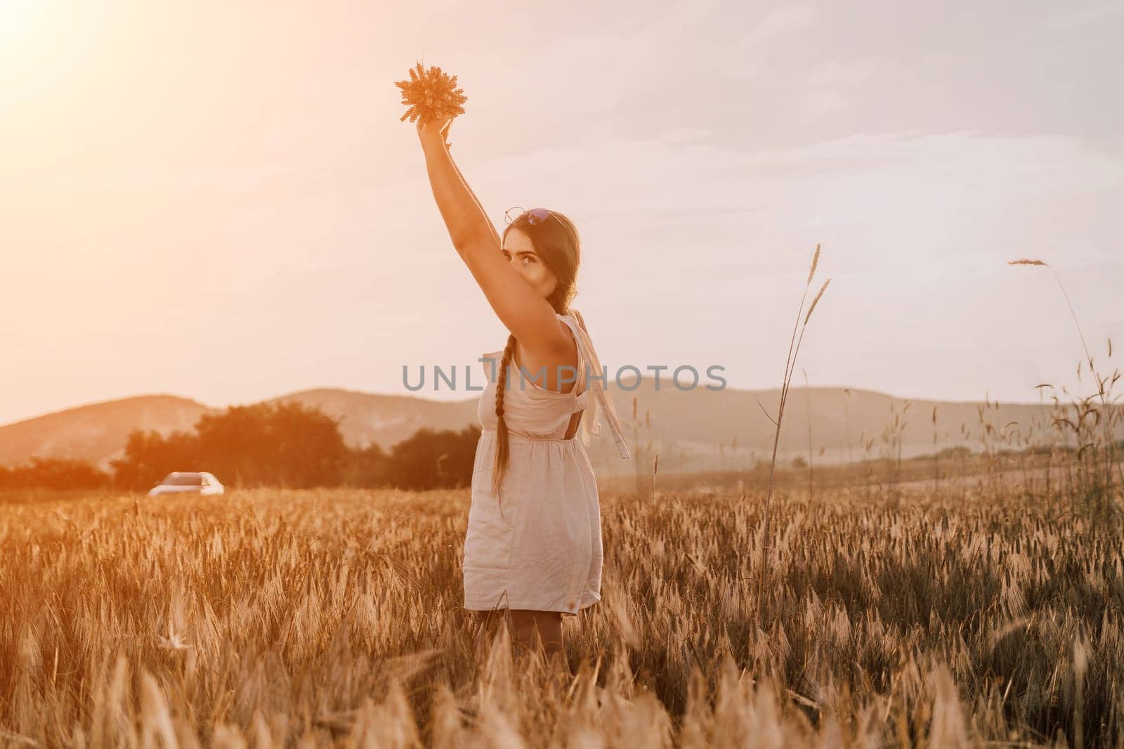 A woman is holding a bunch of wheat in her arms. The wheat is dry and brown, and the woman is wearing a white dress. The scene is set in a field, and the woman is posing for a photo