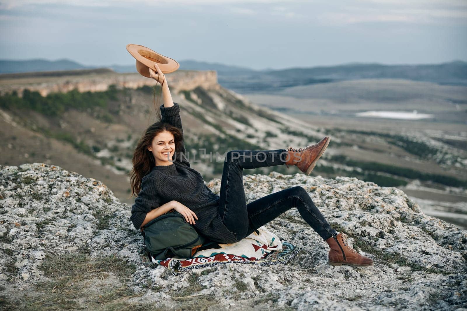 Serene woman relaxing on mountain peak with hat and bag, enjoying tranquil nature view by Vichizh