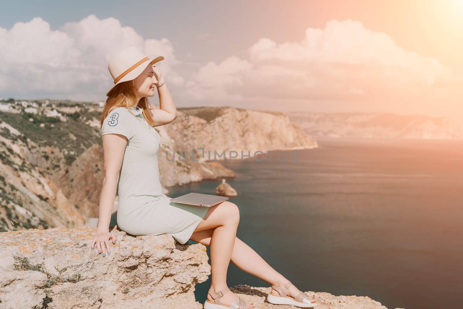 Digital nomad, woman in the hat, a business woman with a laptop sits on the rocks by the sea during sunset, makes a business transaction online from a distance. Freelance, remote work on vacation.