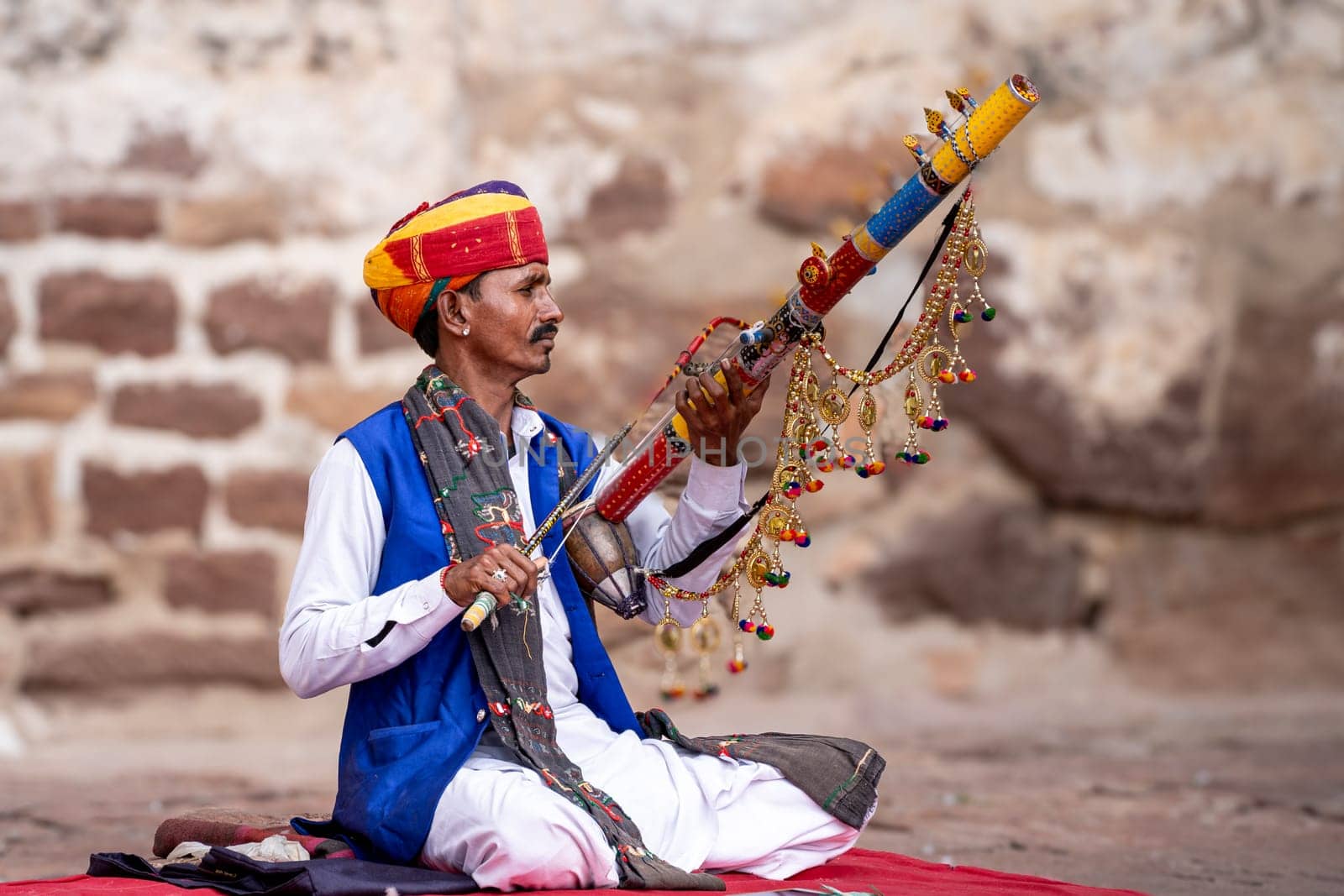 Jodhpur, Rajasthan, India - 26th Dec 2023: Rajasthani Indian musician sitting cross legged in colorful turban and white kurta pyjama local clothing in front of a brick wall at a mahal palace
