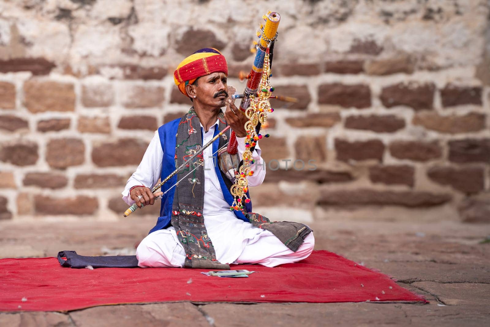 Rajasthani Indian musician sitting cross legged in colorful turban and white kurta pyjama local clothing in front of a brick wall at a mahal palace by Shalinimathur