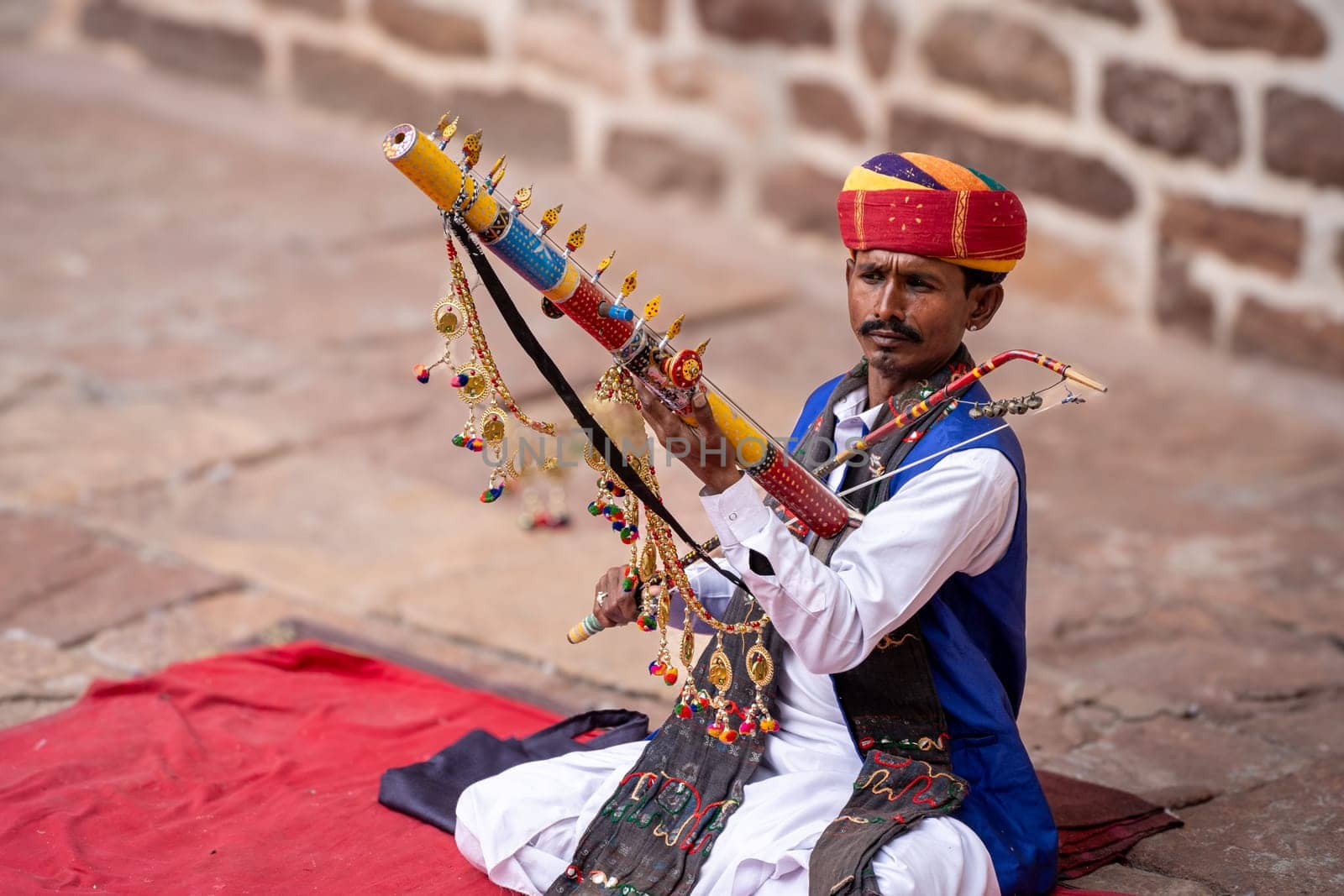 Rajasthani Indian musician sitting cross legged in colorful turban and white kurta pyjama local clothing in front of a brick wall at a mahal palace by Shalinimathur