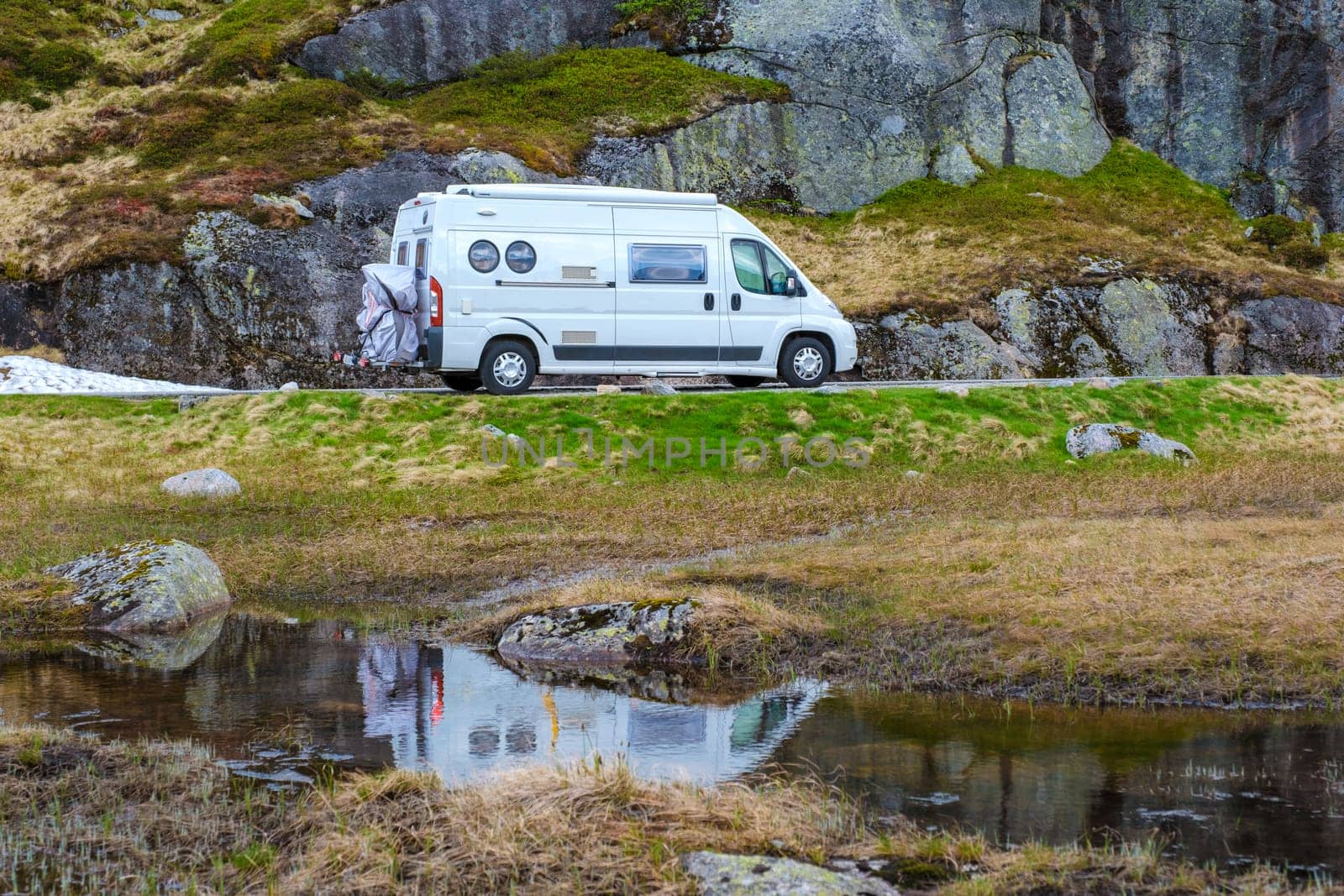 A white campervan drives along a winding road in Norway, its reflection shimmering in a nearby pool of water.