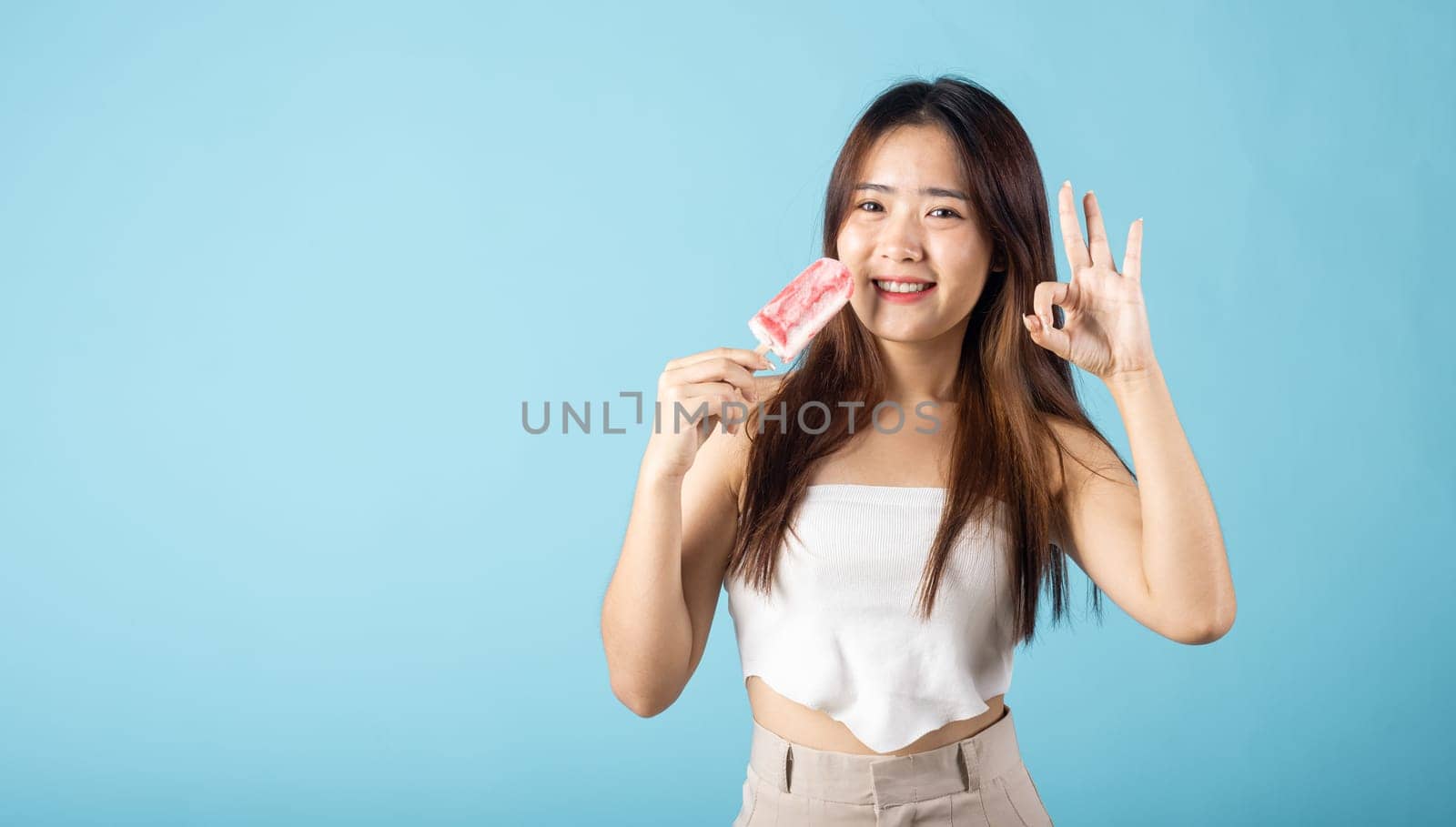 Sweet tasty frozen dessert on summer time. Happy Asian young beautiful woman holding delicious ice cream wood stick mixed fruit flavor and show OK sign, studio shot isolated on blue background