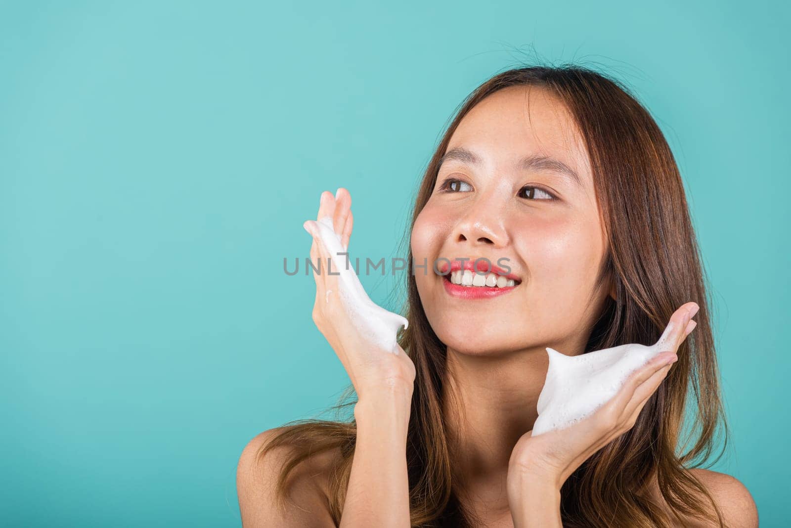 Portrait Asian young woman hands are adorned with white foam mousse studio shot isolated on blue background, beautiful female holds a cleansing foam near her face, Cosmetics beauty skincare