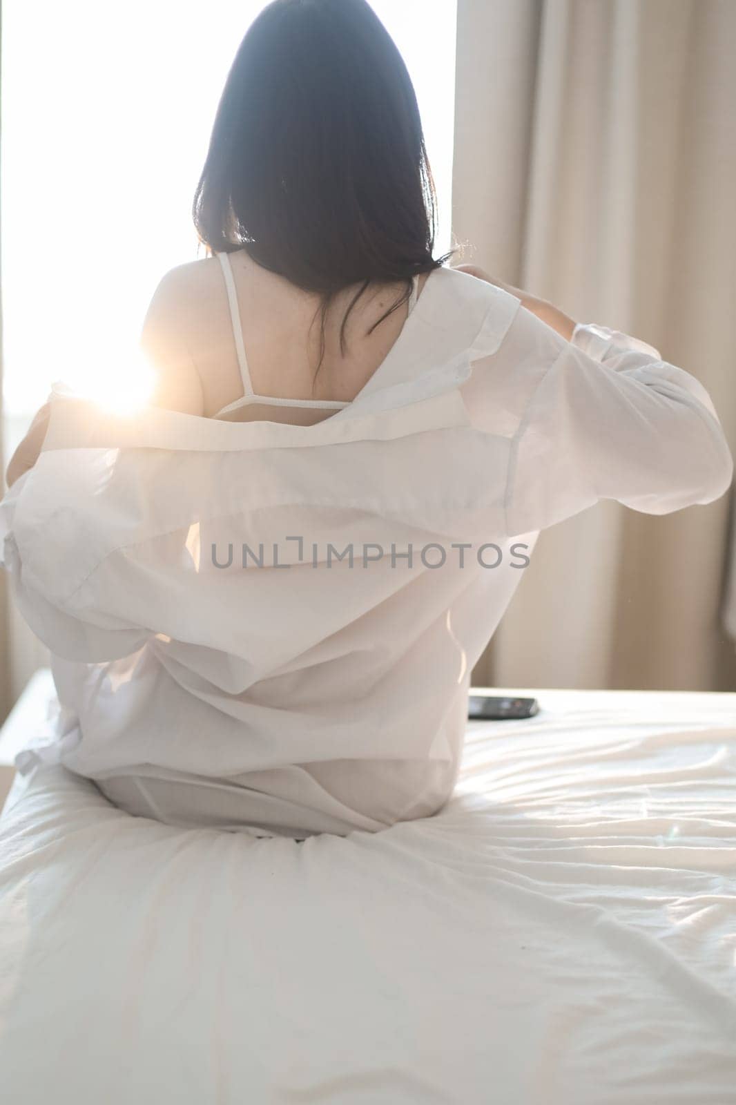 Young woman in lingerie and white shirt sit on bed indoors at home stretching in the morning after waking up, back view. Relaxing , Cozy, good morning, comfort, intimacy concept.