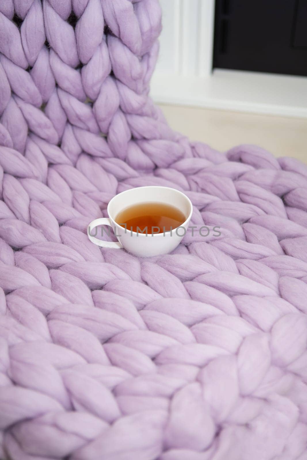 The concept of coziness and comfort is a pink-lavender knitted blanket with a white cup of hot tea on it. Place for inscription, vertical photo