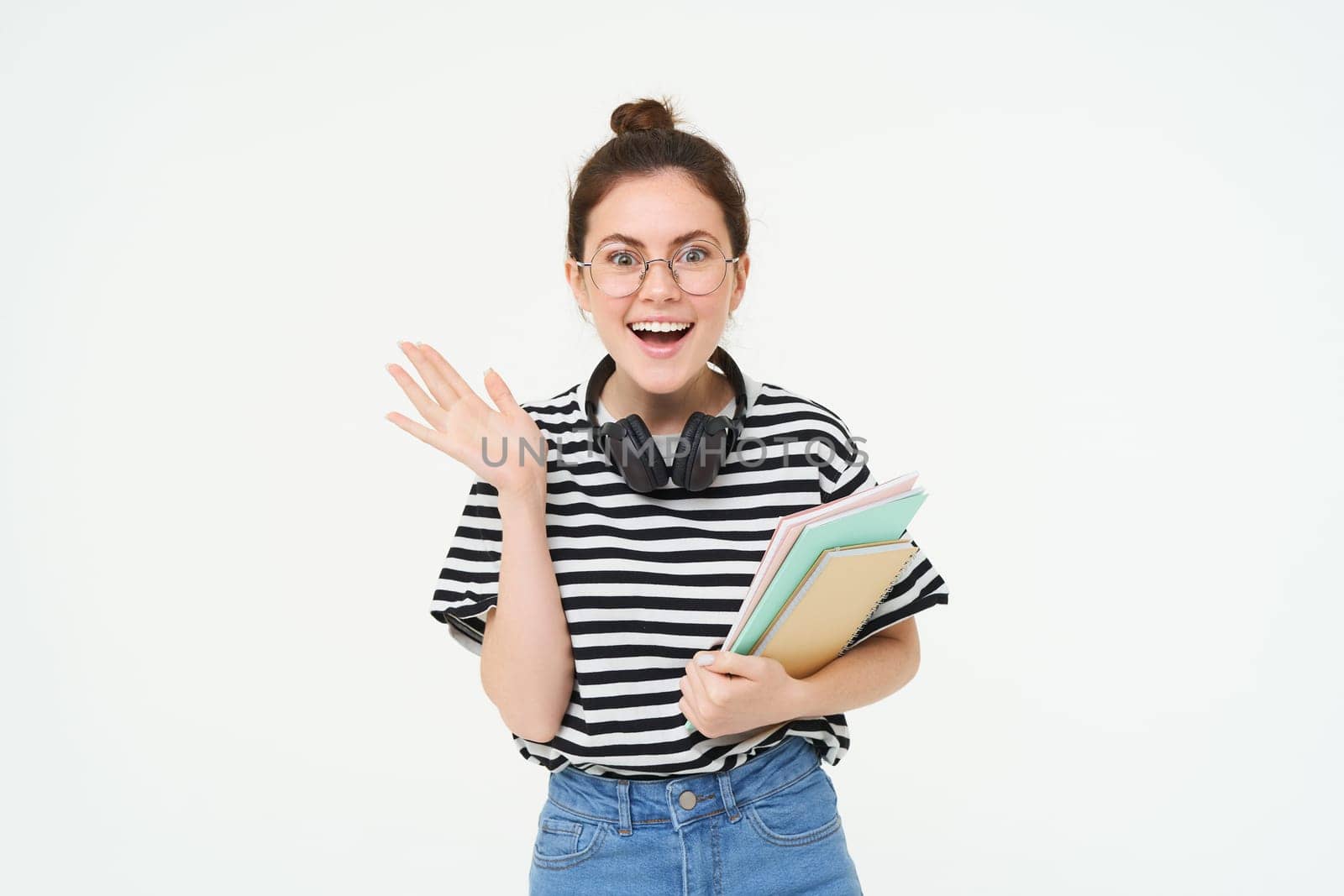 Portrait of excited young woman in glasses, looks surprised and happy. Student standing on white background with books and notebooks.