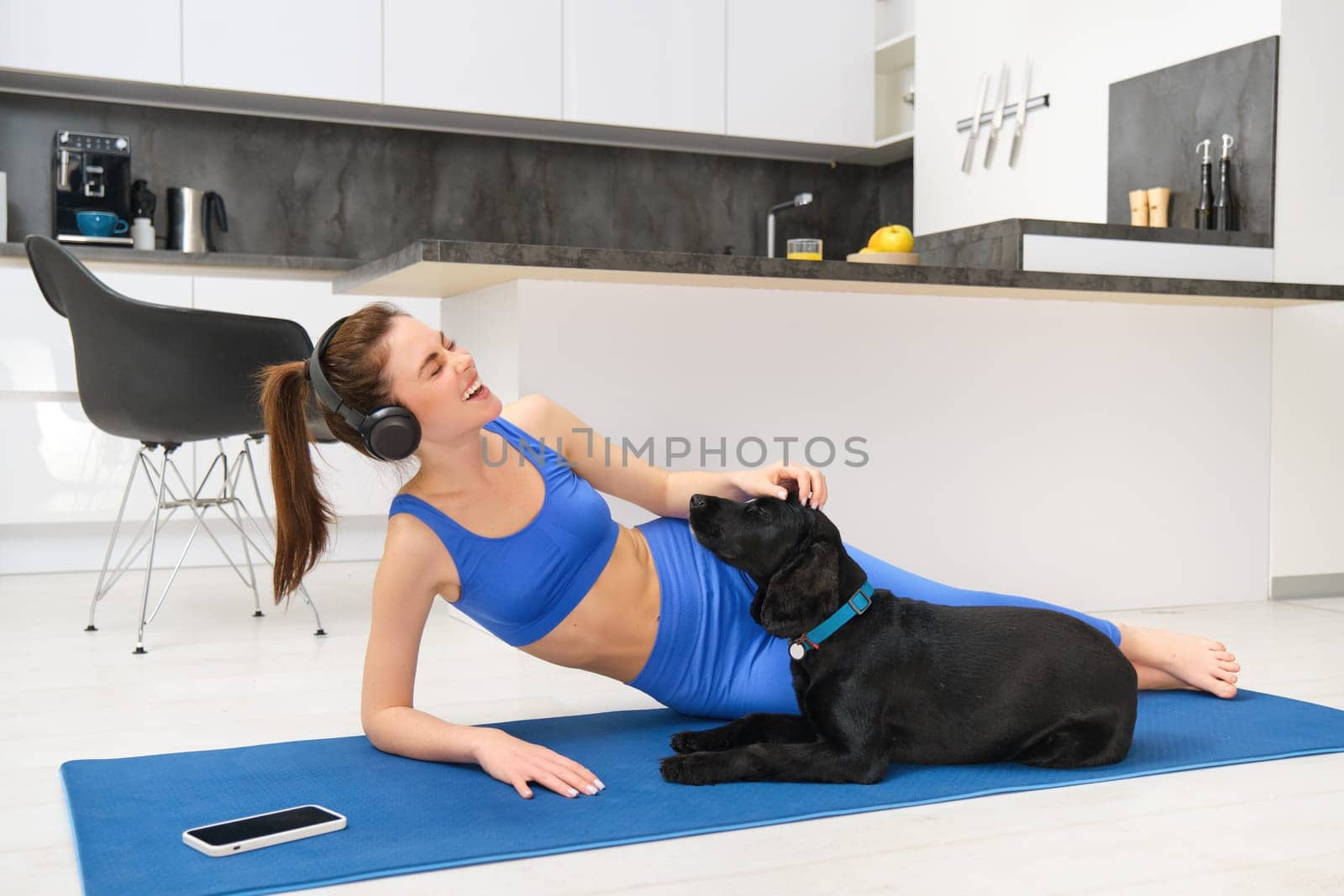 Lifestyle and fitness concept. Happy young woman doing aerobics while her dog wants to play, laughing and smiling, laying on yoga mat.