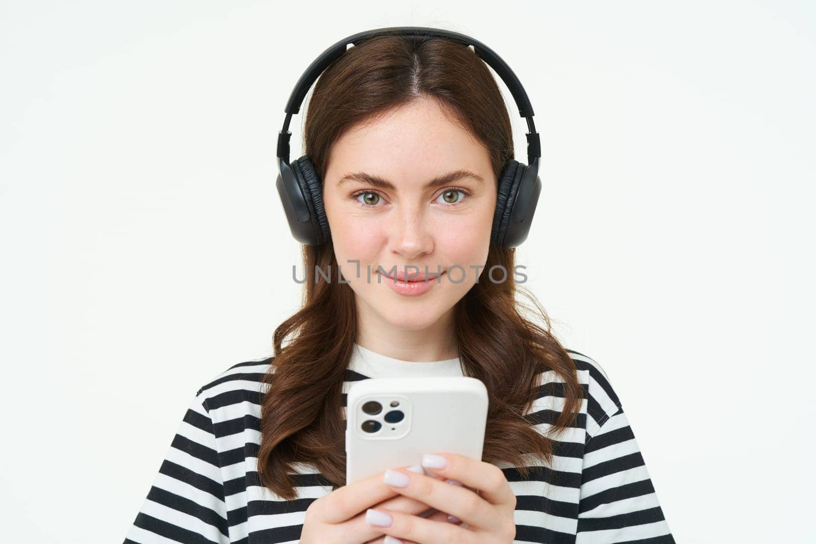 Image of brunette young woman, smiling, listening to music in headphones, watching videos on mobile phone app, holding smartphone, standing over white background by Benzoix