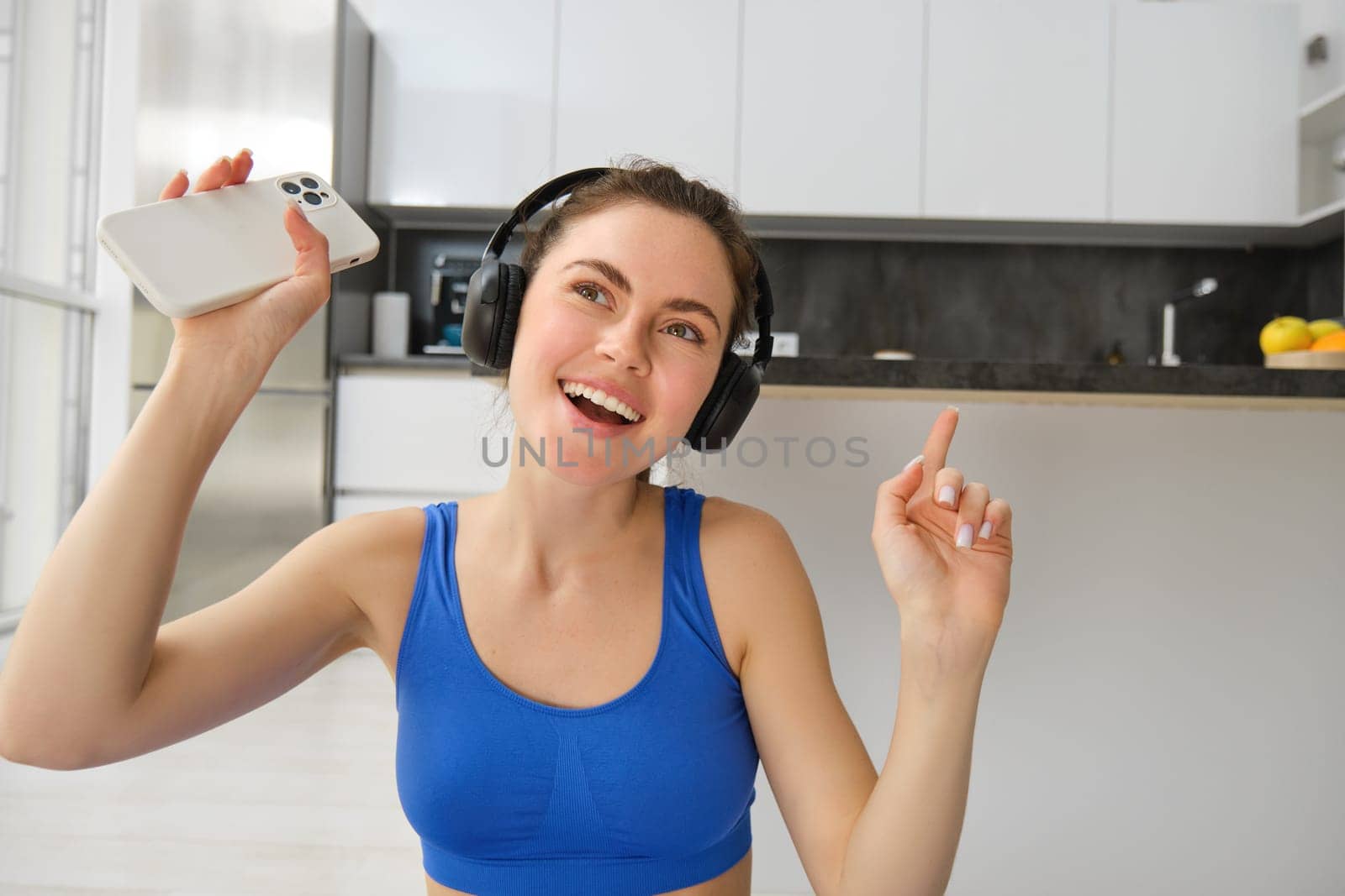 Close up portrait of happy fitness girl, listens music in wireless headphones, holds smartphone, dances and workout from home.