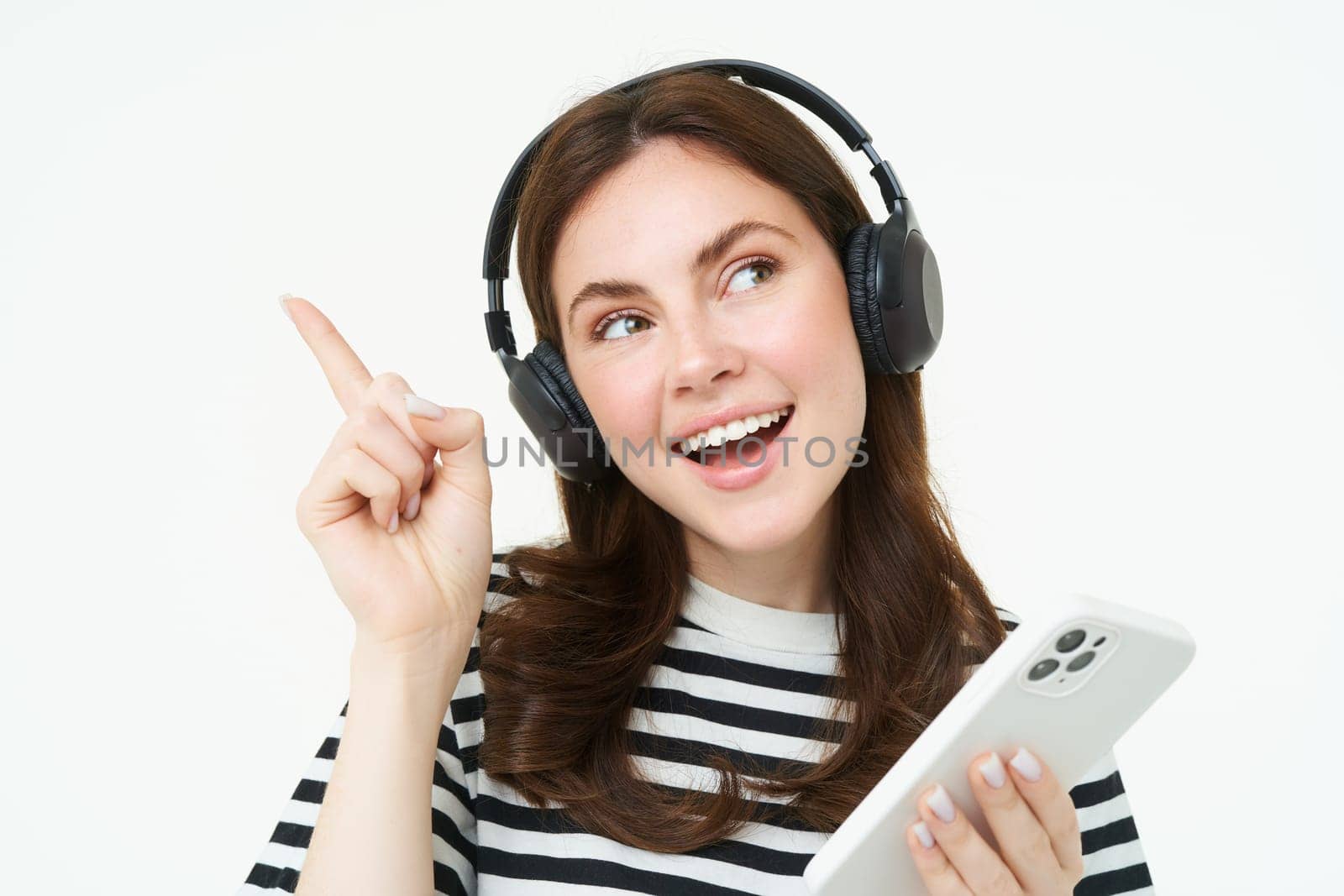 Happy beautiful woman, listening music in wireless headphones, holding mobile phone, pointing left at copy space, showing advertisement, white background.