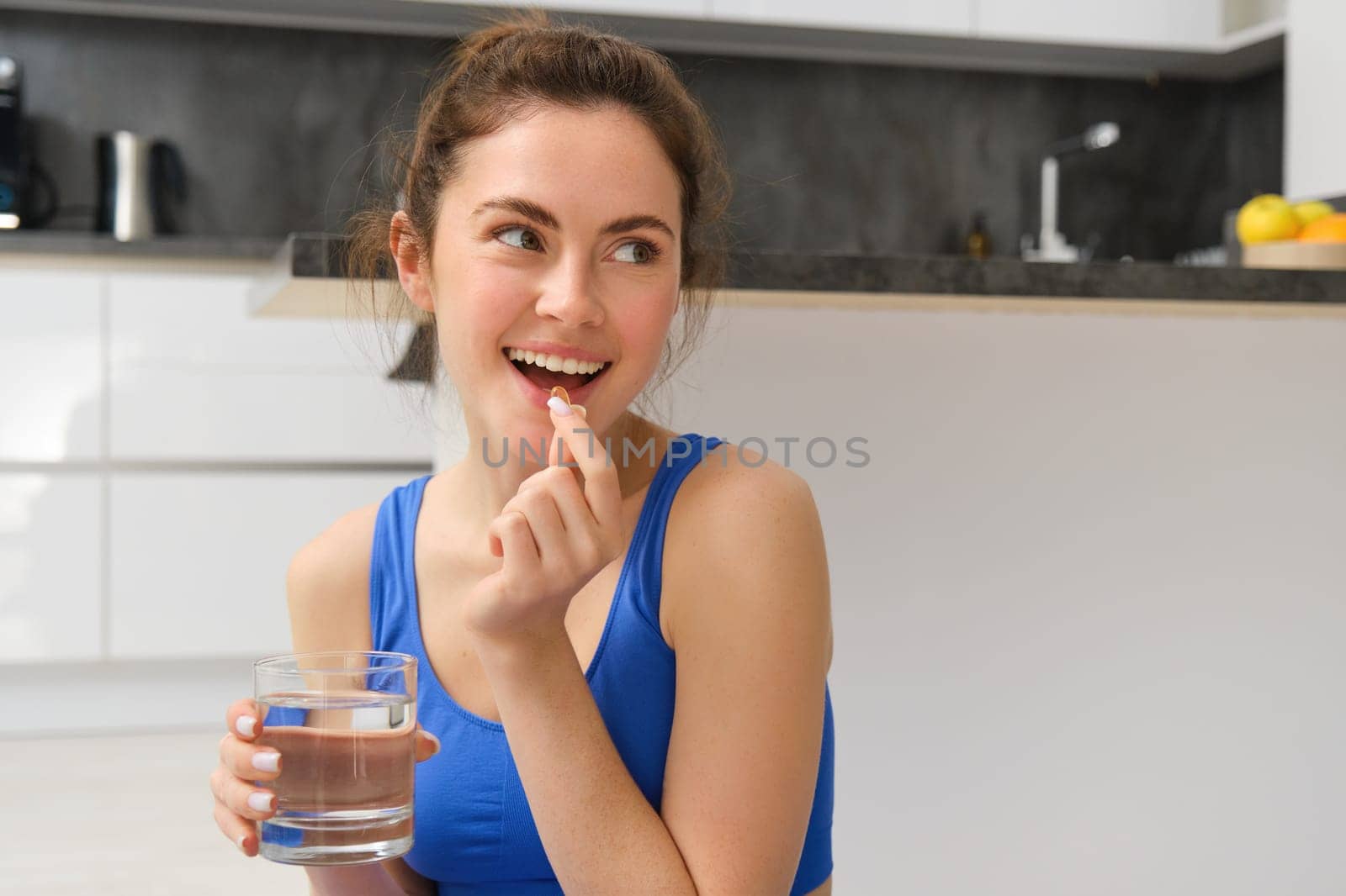 Close up portrait of brunette fitness woman, taking dietary supplements, vitamins and glass of water after workout at home.