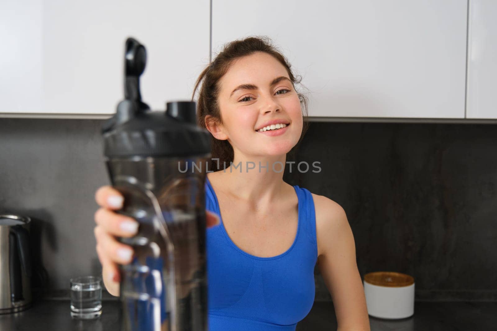 Image of fit and healthy, smiling young woman, offering bottle of water, give you a drink after fitness workout, standing in kitchen.