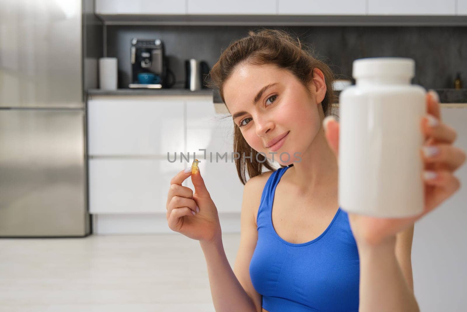 Close up portrait of young woman, fitness instructor showing bottle of vitamins, taking buds supplementary, dietary pill, sitting at home, doing workout exercises by Benzoix