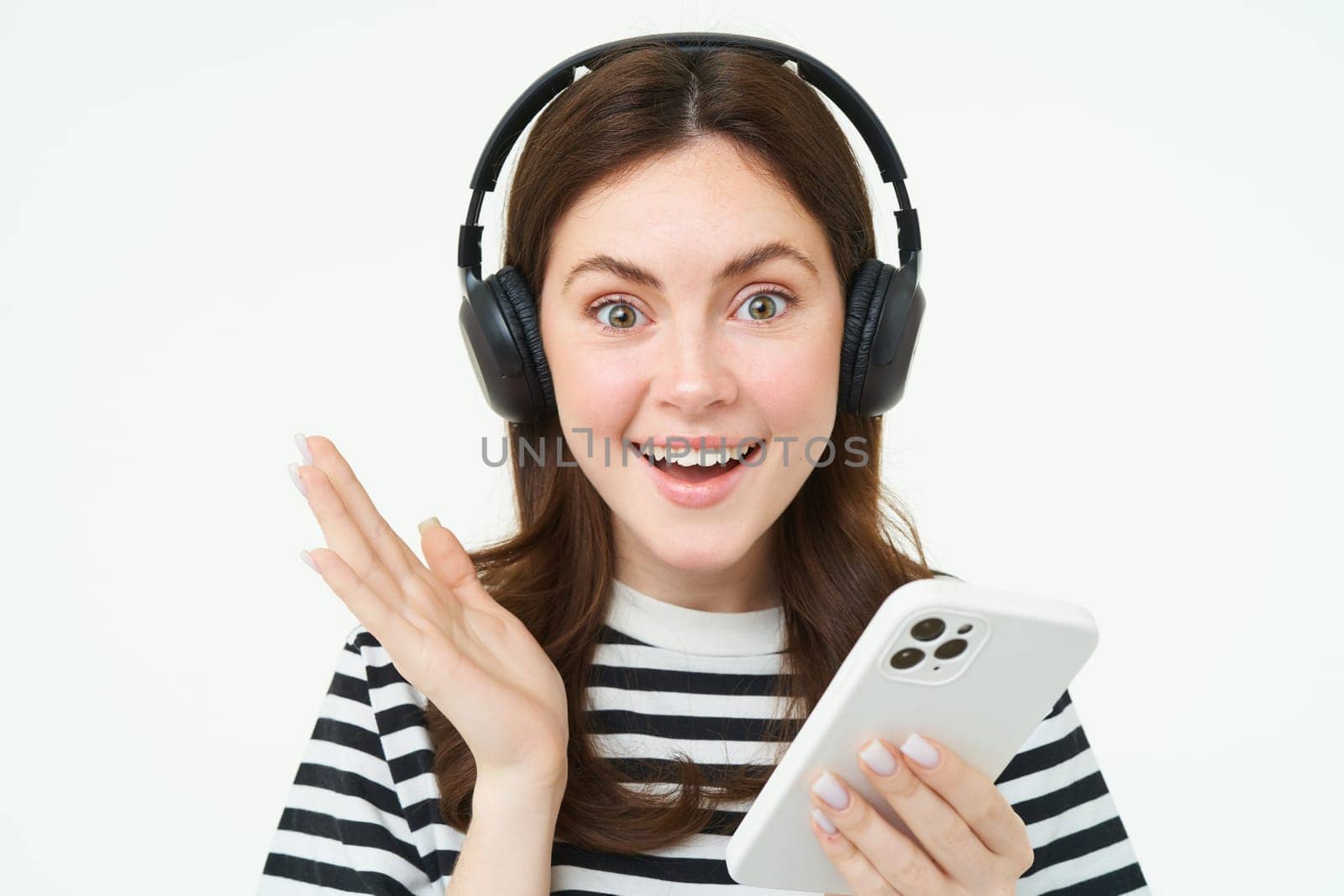 Cheerful, beautiful young woman using smartphone app, celebrating, looking happy while playing with mobile phone, listening music in wireless headphones by Benzoix