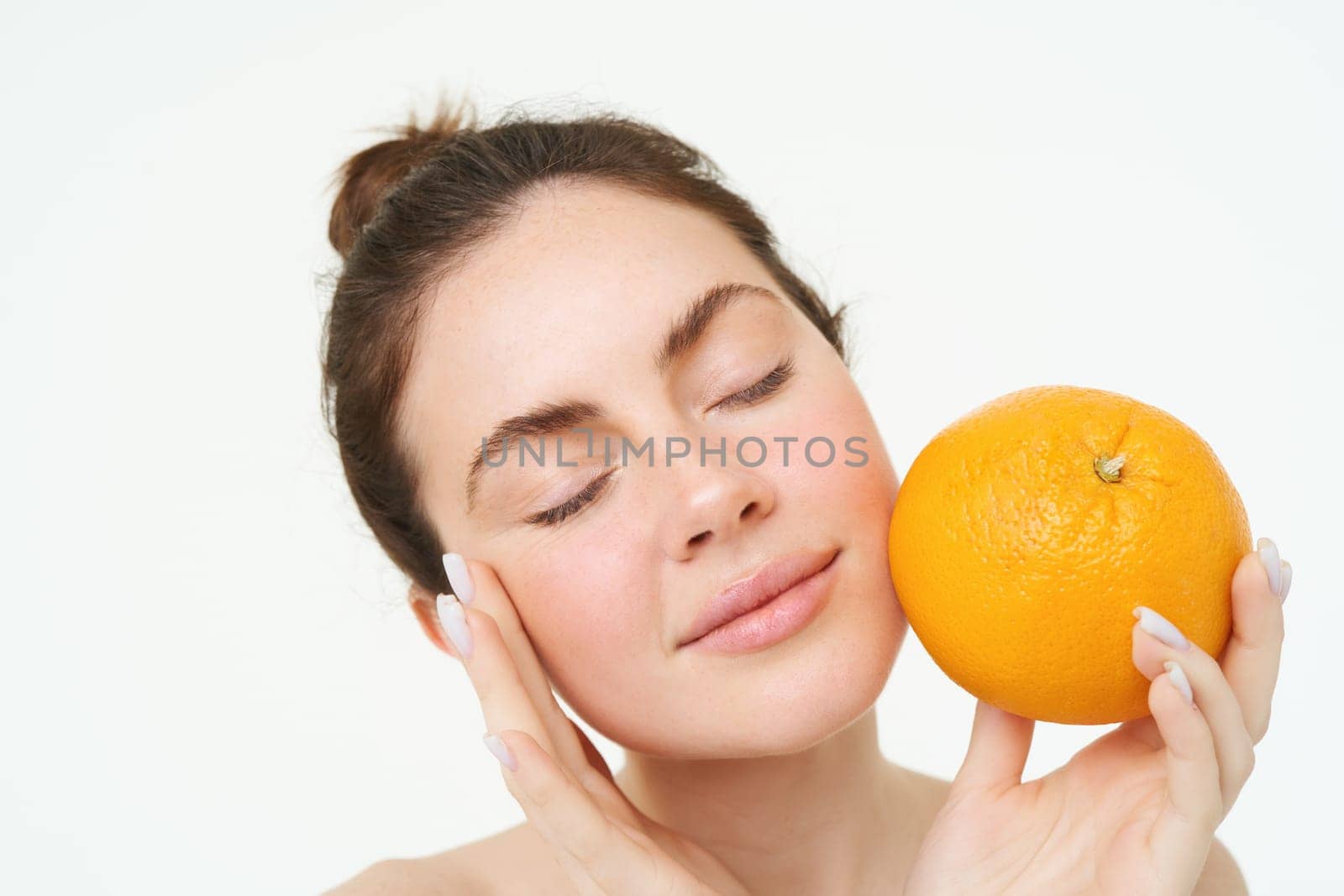 Portrait of young woman holding orange near face without blemishes, smiling, concept of vitamins, nourishment and organic facial treatments, white background by Benzoix