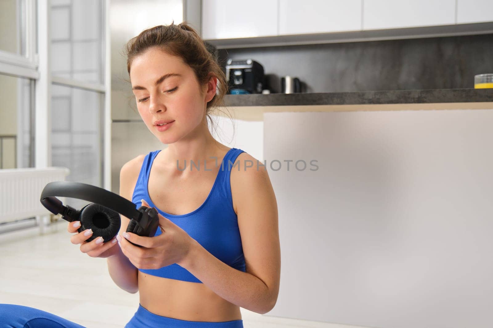 Image of young fitness girl, wearing blue tracksuit, puts on wireless headphones to workout with music, does yoga at home by Benzoix