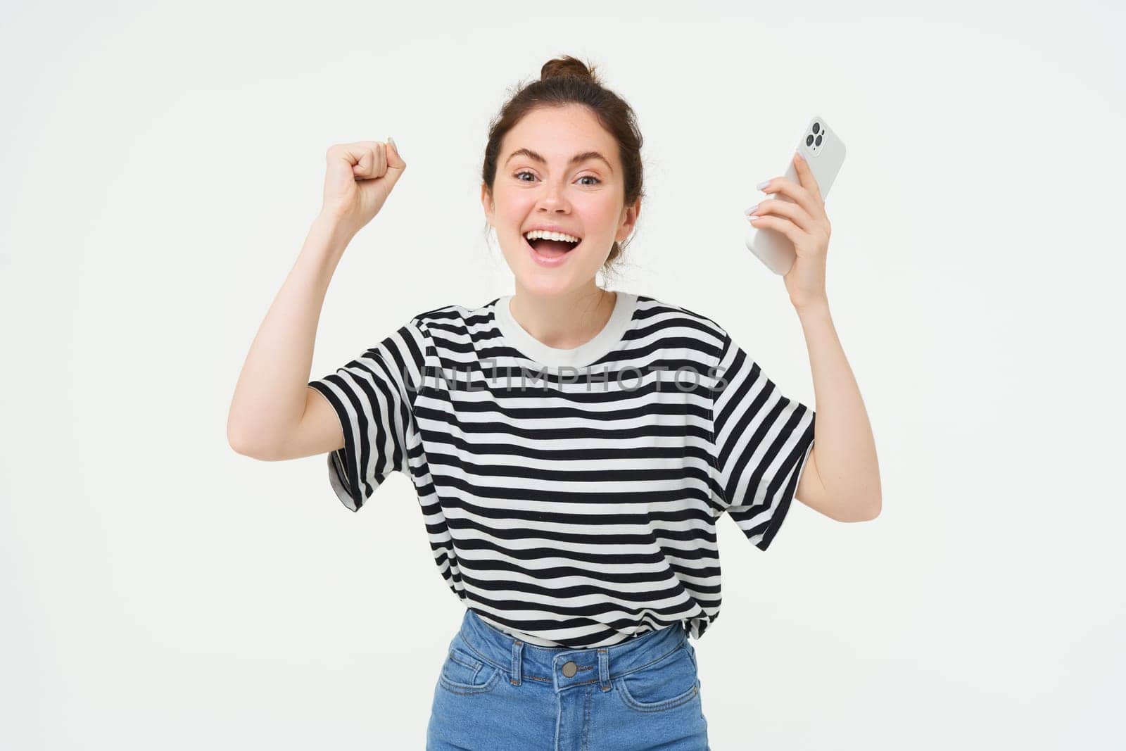 Portrait of happy woman laughing, holding telephone, using smartphone and chanting, rooting for someone, holding hands up, isolated over white background by Benzoix