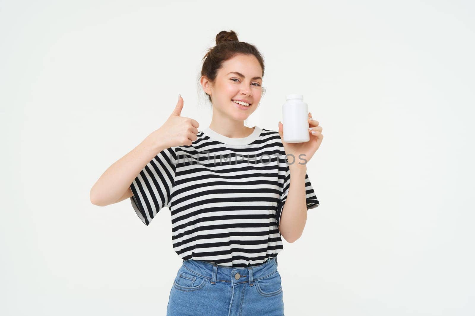 Portrait of beautiful, smiling brunette woman, shows thumbs up and bottle of vitamins, recommends dietary supplements, isolated against white background.