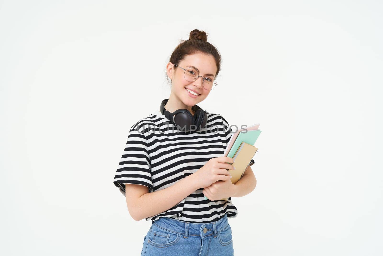 Education concept. Smiling brunette girl, student in casual clothes, holds her books, study material, wears headphones over neck, looks confident and happy, isolated over white background.