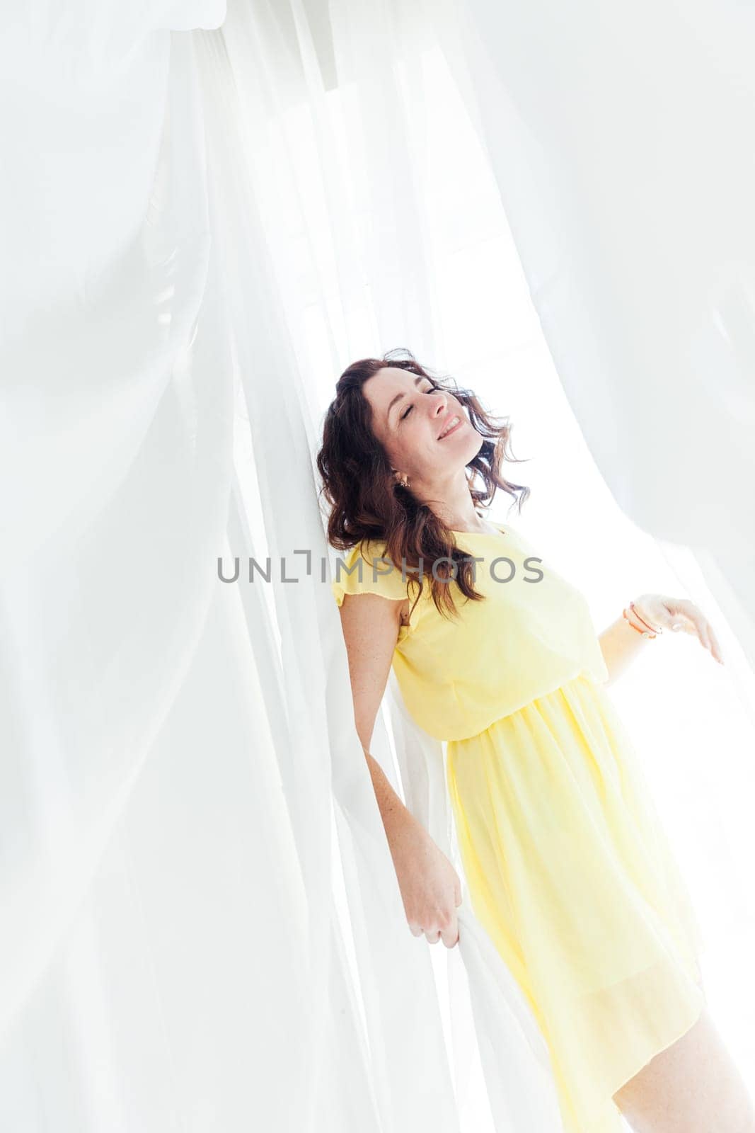 beautiful brunette woman in yellow dress stands at the white curtains of tulle fabrics in a bright room