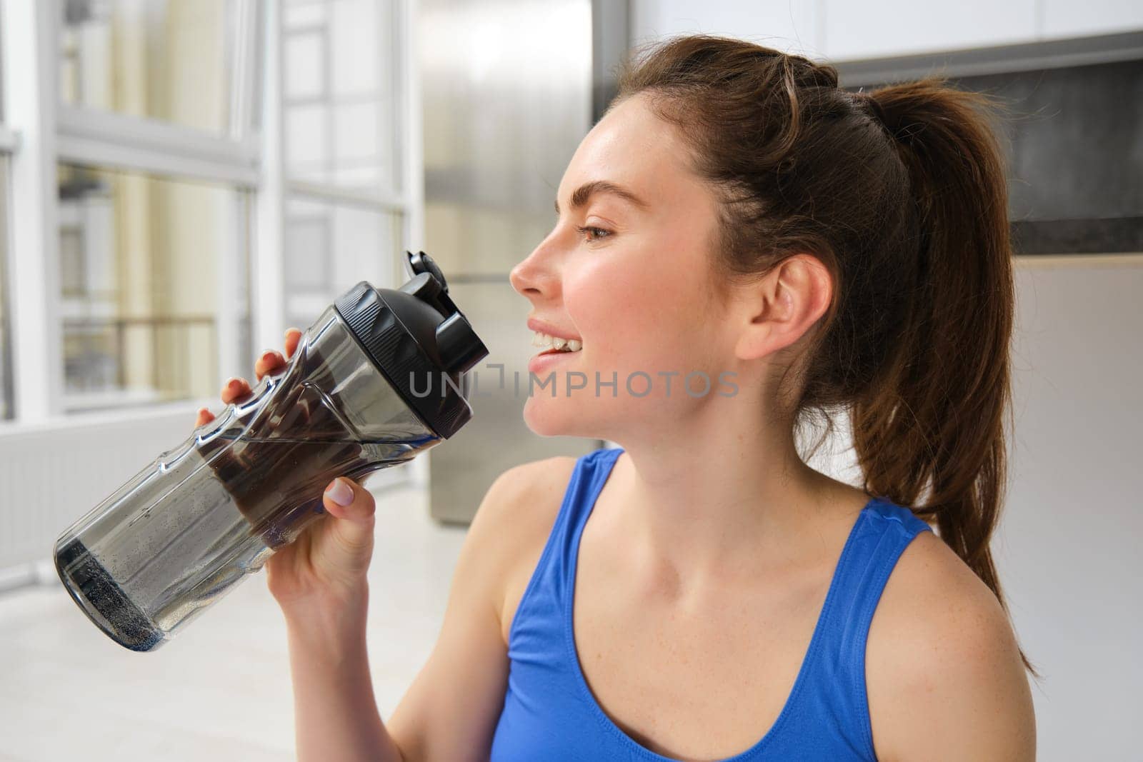 Beautiful young sportswoman drinks water, stays hydrated during workout training at home, doing fitness exercises in living room.