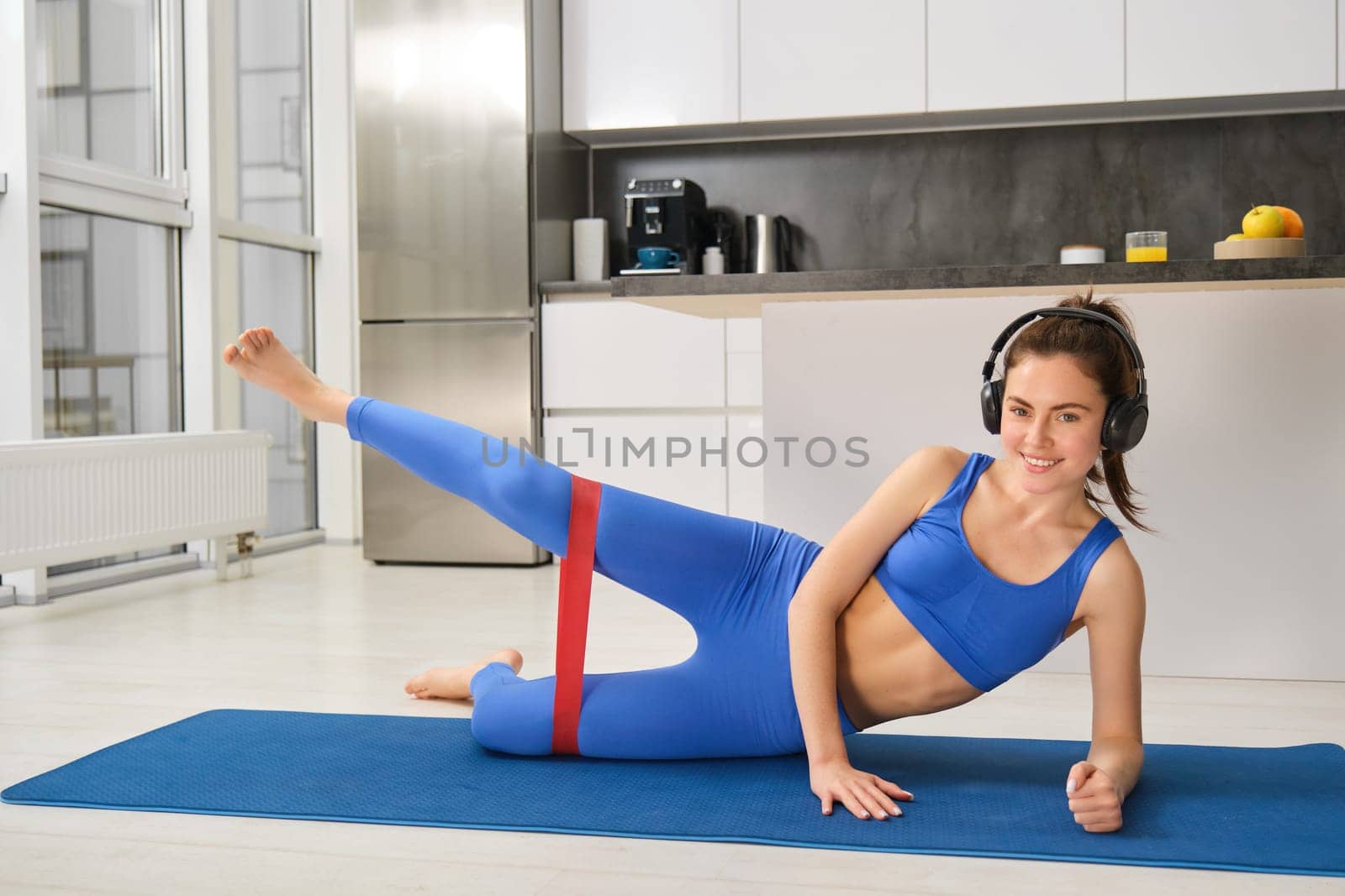 Workout and fitness concept. Woman in headphones does exercises at home, stretching rubber resistance band with legs, laying on yoga mat in living room, lifting her foot up by Benzoix