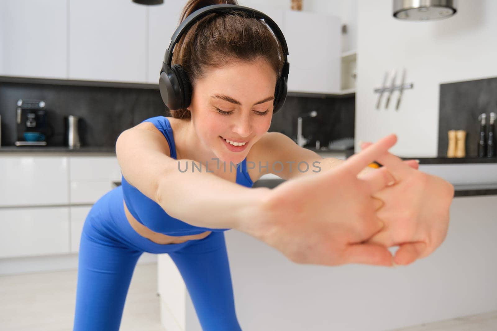 Close up portrait of fit and healthy woman, smiling while doing workout exercises, listening music in wireless headphones, stretching arms, aerobics training at home, wearing blue activewear.