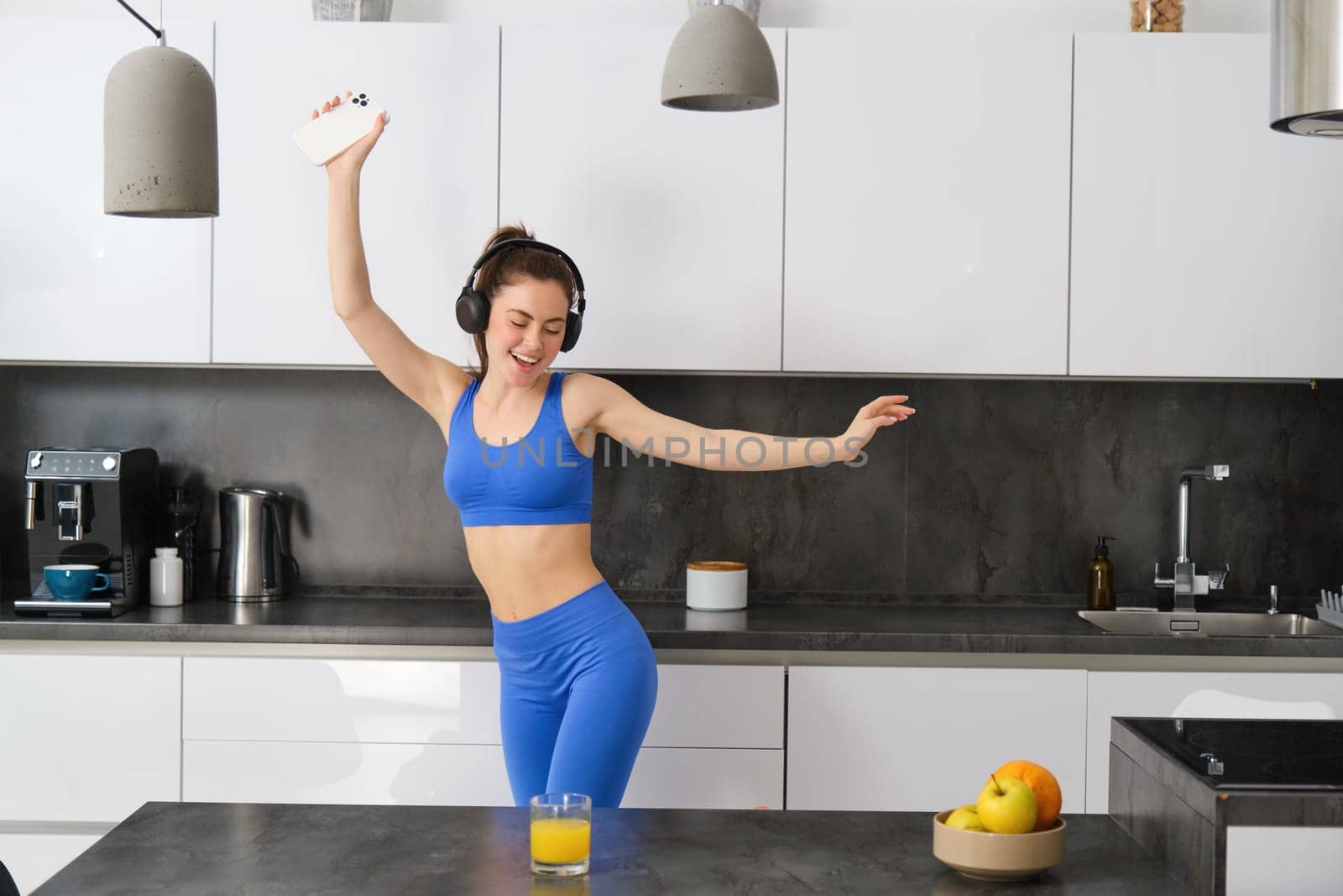 Portrait of beautiful sports woman, wearing activewear, dancing in kitchen with headphones, listening music and having fun, feeling energy after workout session by Benzoix