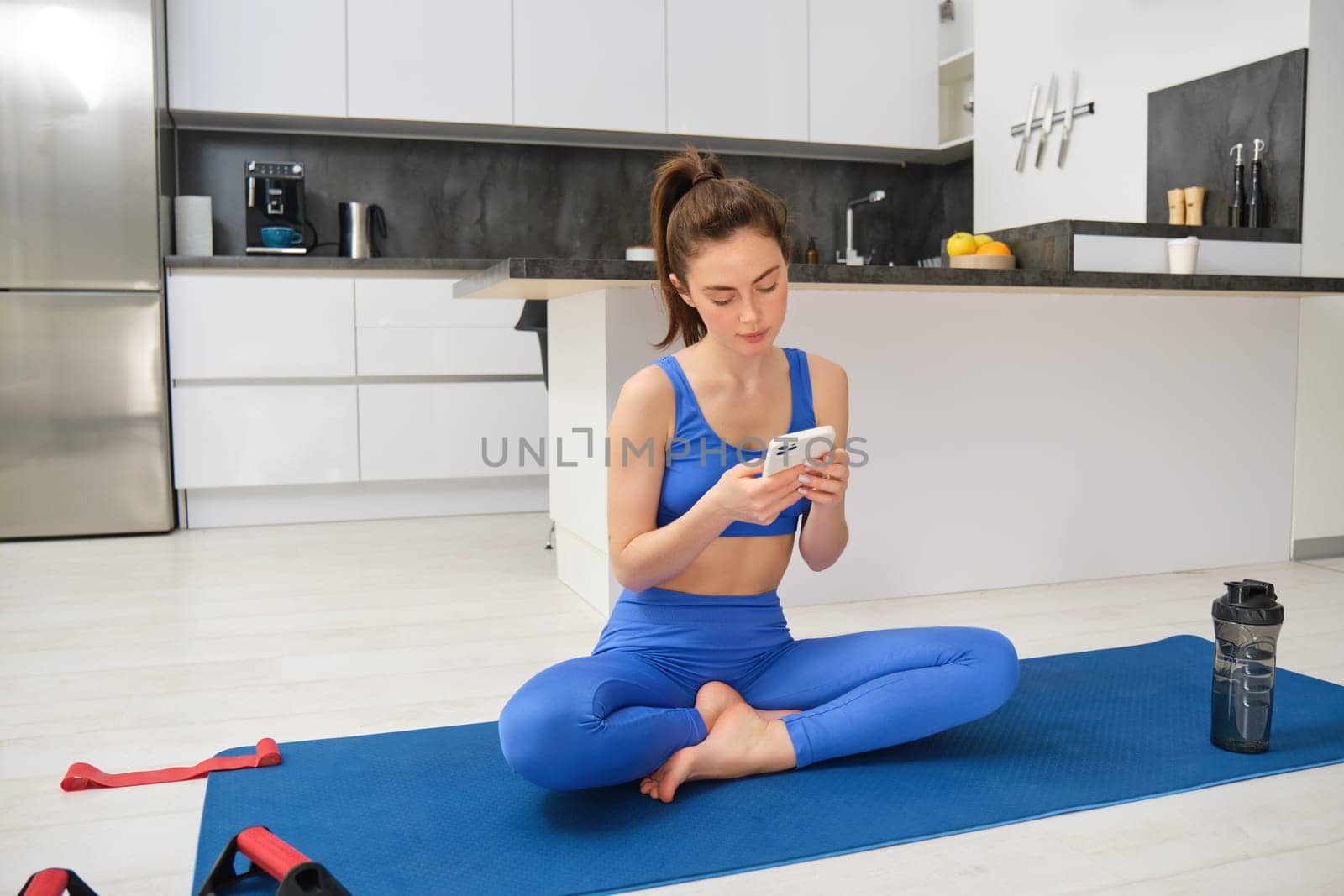 Portrait of sportswoman using sport app while doing workout, yoga exercises at home, sits on mat with water bottle, smiling and looking happy by Benzoix