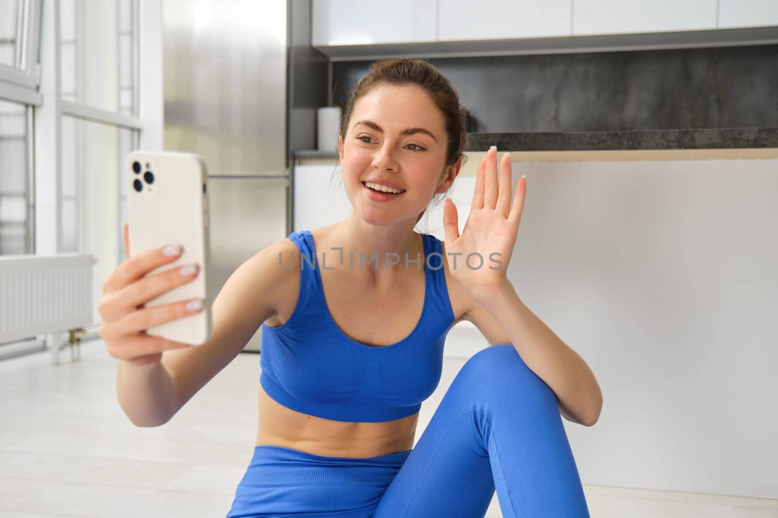 Image of young woman doing workout, takes selfie, records online video. Fitness instructor records her training on phone, says hello at smartphone camera and waves hand at it by Benzoix