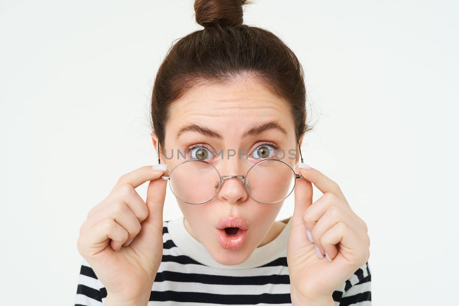 Close up portrait of surprised girl, takes off glasses, reads amazing news, impressed by something, stands over white background.