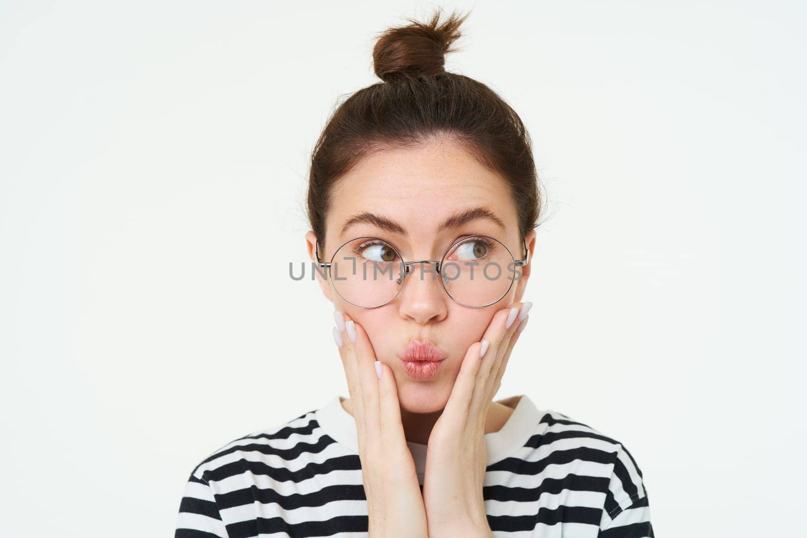 Close up portrait of cute girl in glasses, holds hands on face and puckers lips, looks aside, makes silly face expression, stands over white background.