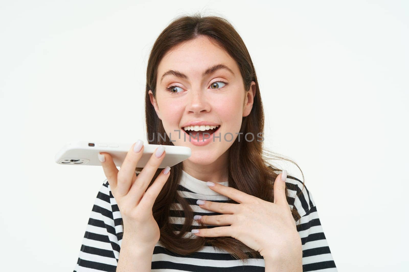 Image of cute brunette woman talking into speakerphone, holding mobile phone near mouth, records her voice, sends a voicemessage, using online translator app, white background.