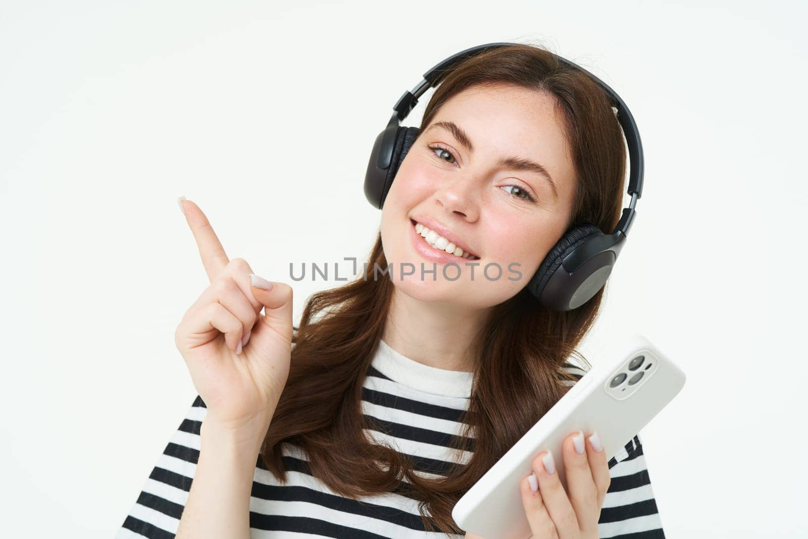Portrait of smiling student, girl in headphones, holding mobile phone, pointing left, showing advertisement, store offer, white background.
