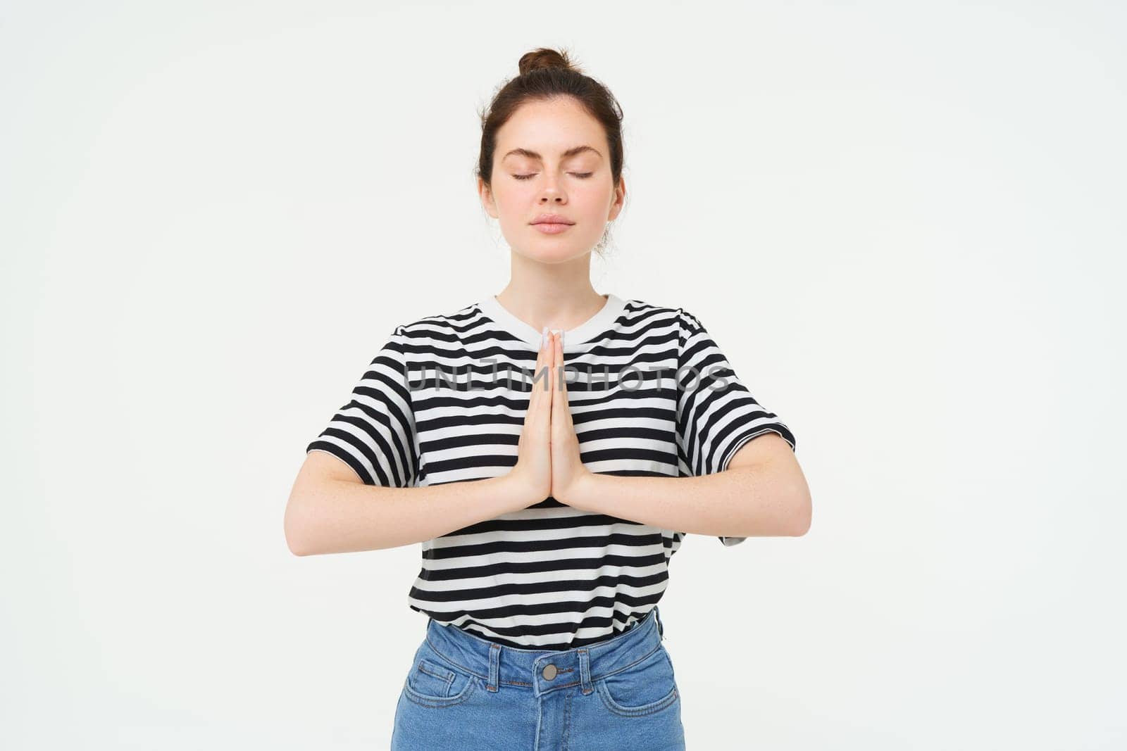 Portrait of young mindful woman meditating, holds hands clasped together, namaste gesture, practice yoga, standing over white background.