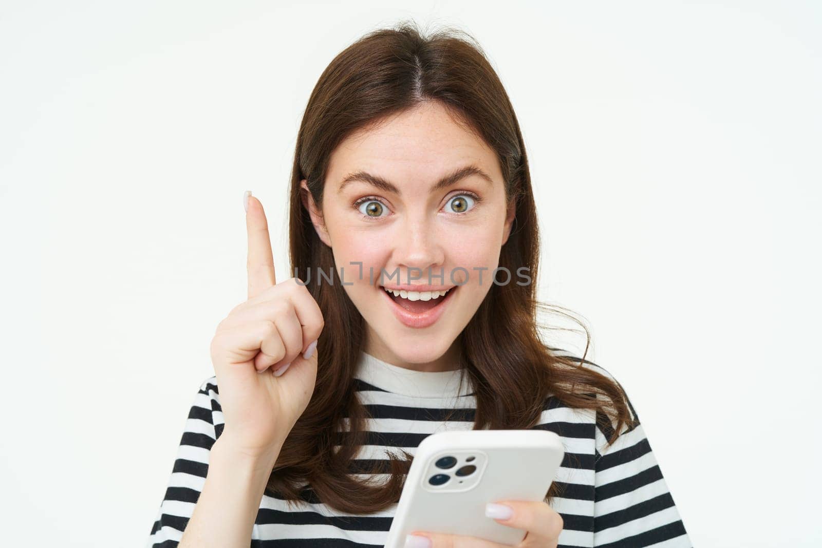 Got an idea. Smiling woman, raises her finger and holds smartphone, suggests something, makes eureka gesture, has a solution, white background.