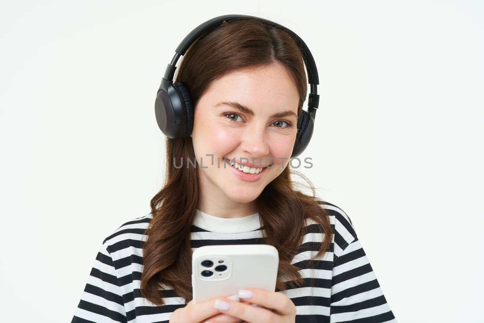 Image of brunette young woman, smiling, listening to music in headphones, watching videos on mobile phone app, holding smartphone, standing over white background.