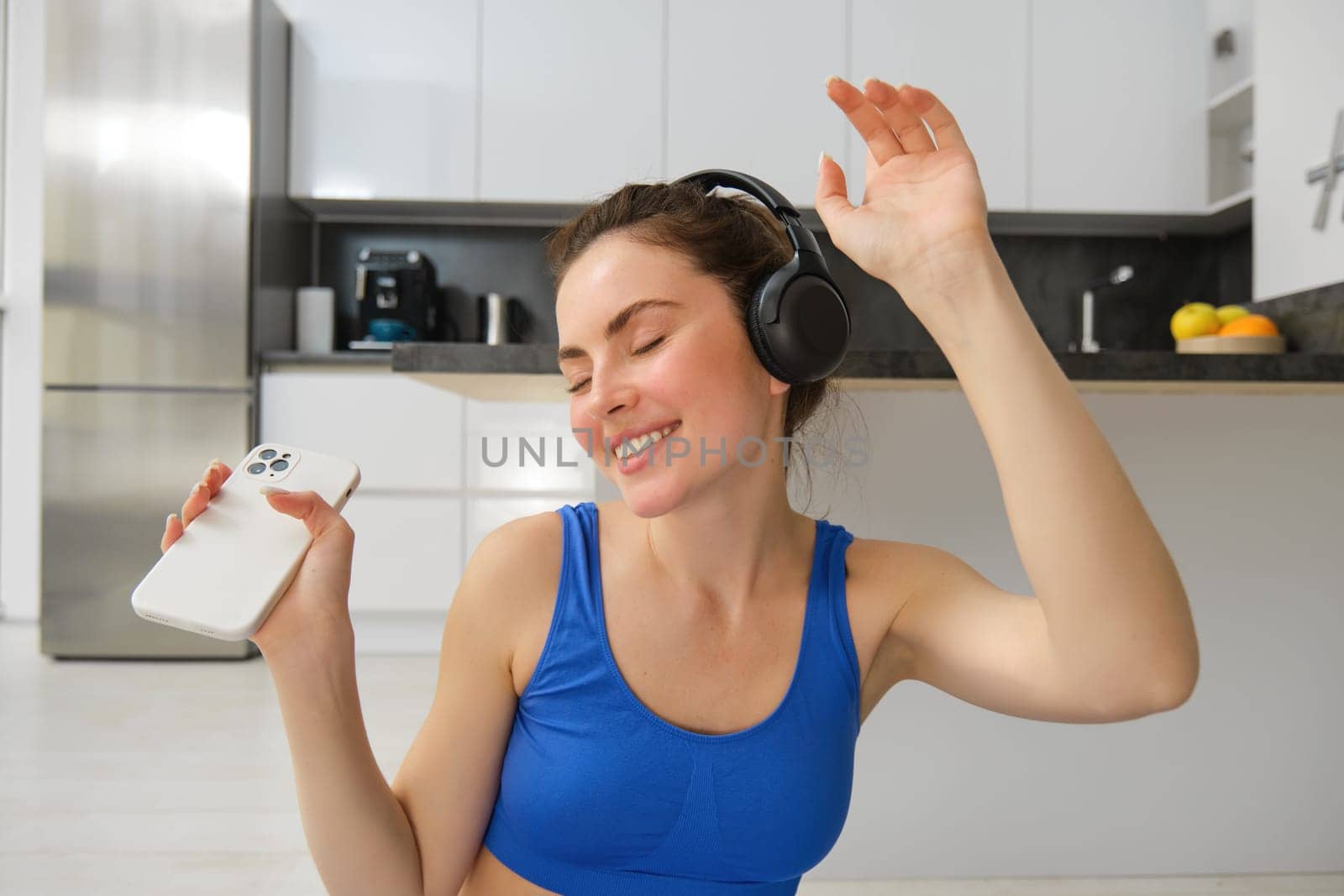 Portrait of fitness instructor, woman with headphones and smartphone, dancing at home, workout inside her house, wearing blue sportsbra by Benzoix