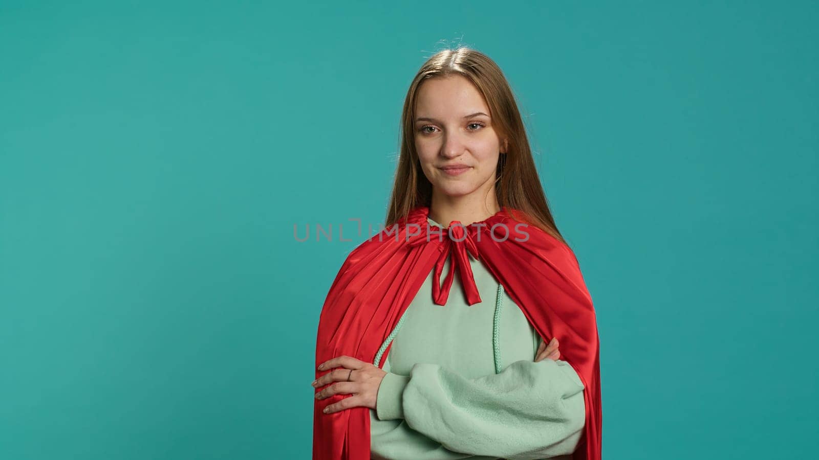 Smiling teenager portraying superhero wearing red cape, isolated over studio background. Portrait of cheerful heroic person posing as hero in costume with mantle, camera A