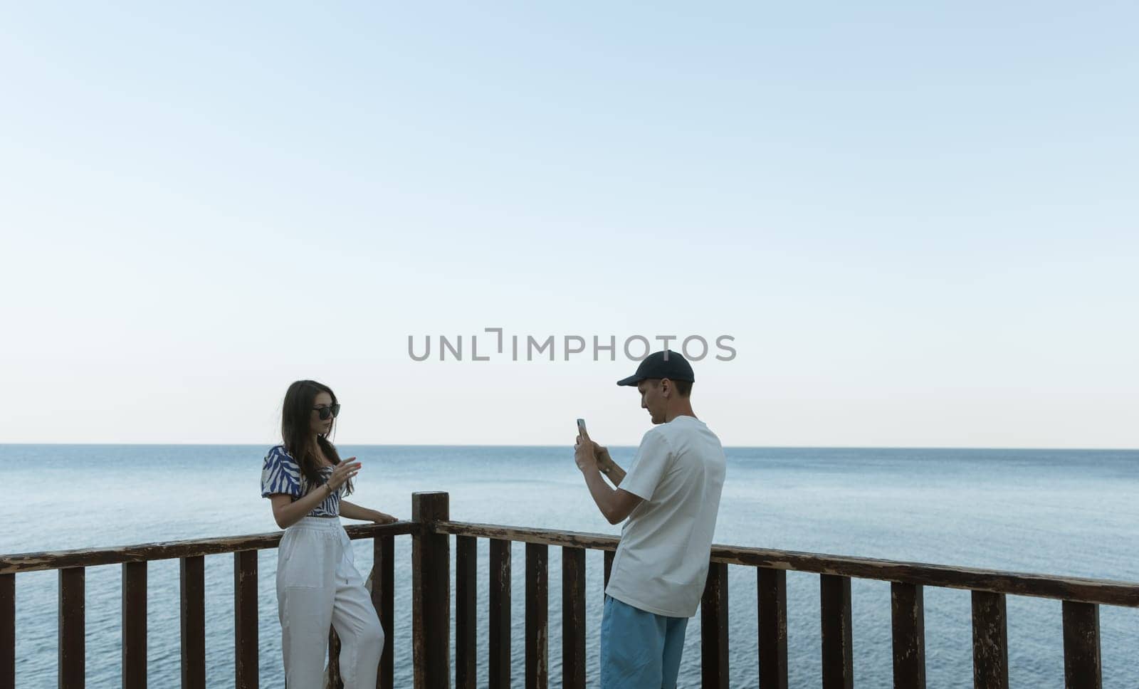 A young guy takes a photo of a girl on his phone by the sea. by Nataliya