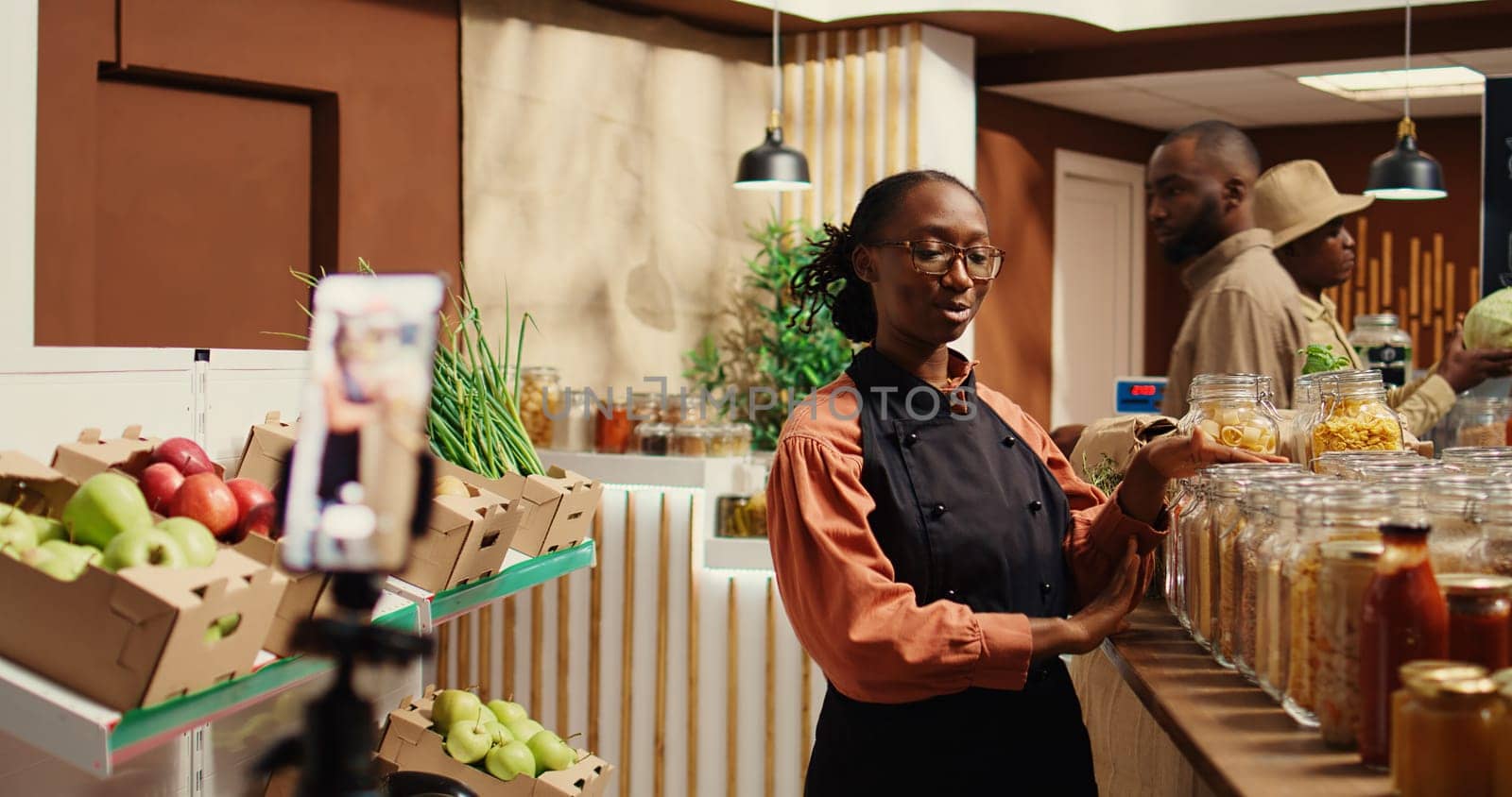 Merchant creating promotional video in local farmers market, talking about natural homemade food sauces and pantry supplies or dressings. African american woman working on marketing. Camera 2.