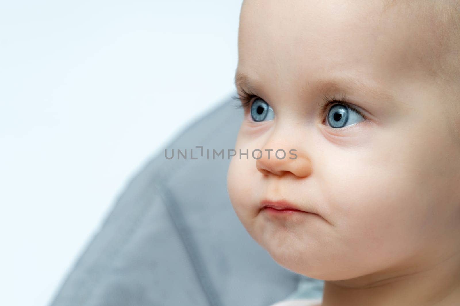 A baby with blue eyes is sitting in a high chair, gazing at the camera with a sweet smile, rosy cheeks, and long eyelashes