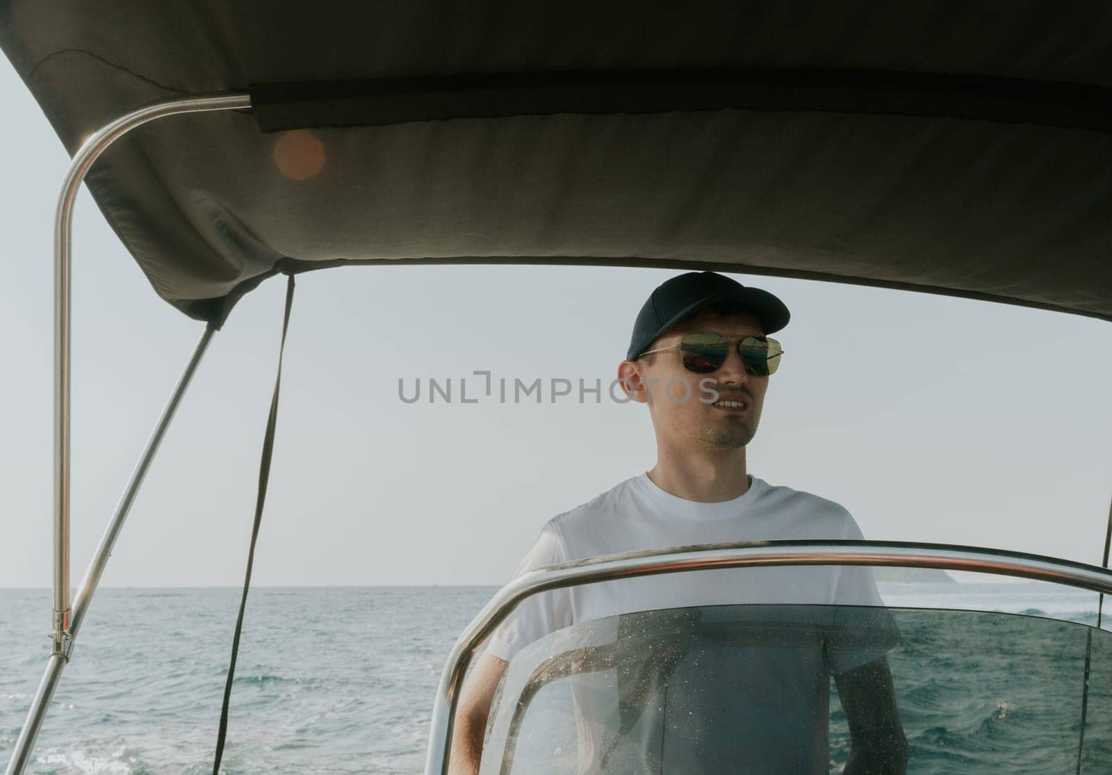 One young Caucasian guy in a cap and sunglasses sails on the sea on a boat, steering the helm and looking forward, on a sunny summer day, close-up side view.