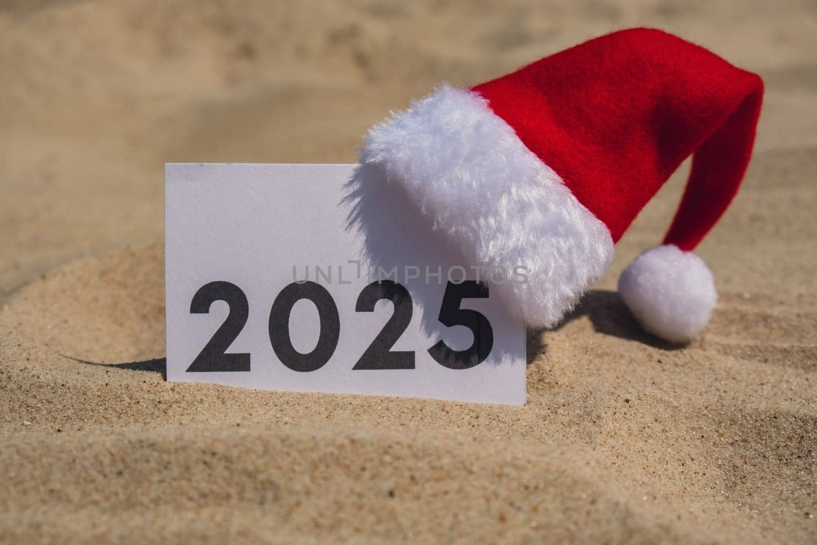 2025 text on paper greeting card on background sandy beach sun coast. Christmas Santa hat New Year New Me decoration. Summer vacation decor. Holiday concept calendar date postcard. Getting away Travel Business concept