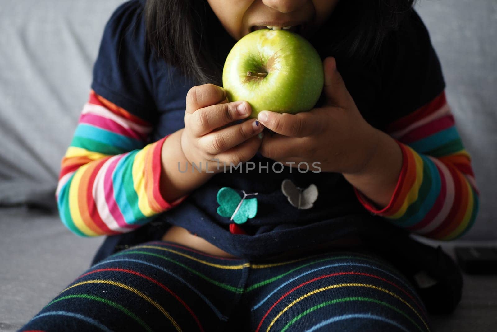 little girl bitting a green apple while sitting on a sofa .