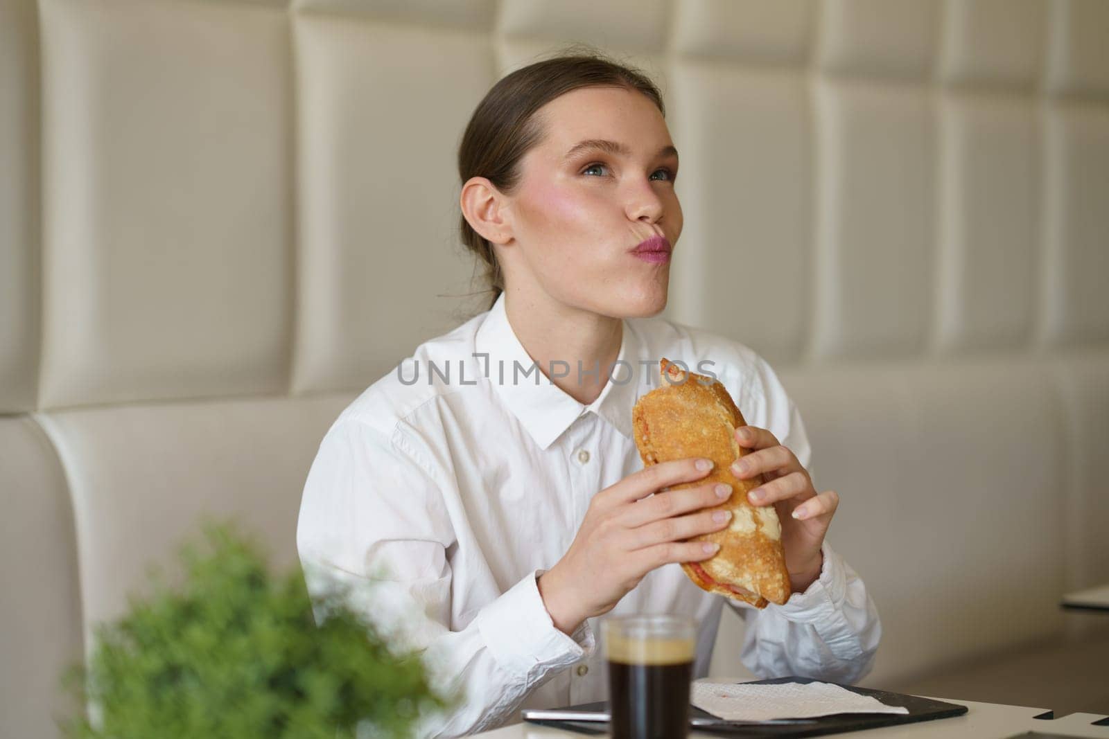 Elegant beauty hungry businesswoman enjoying a delicious sandwich and drinking coffee sitting in a modern cafeteria